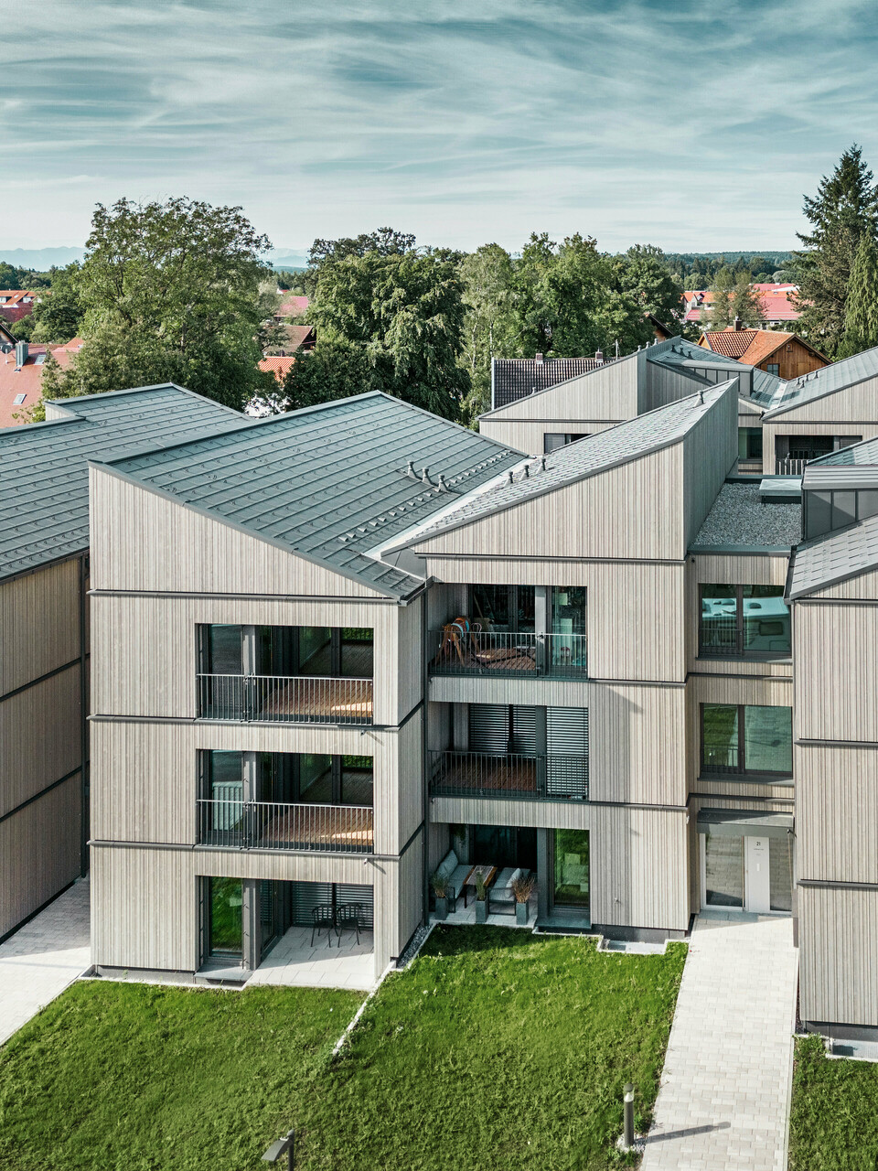 Vista aerea del moderno condominio sul sito Schmucker di Utting am Ammersee, con costruzione ibrida in legno sostenibile ed eleganti facciate in legno. Gli edifici sono dotati di tegole PREFA R.16 in P.10 grigio scuro. Questa architettura innovativa, progettata dallo studio WWA Architekten di Monaco, è caratterizzata da linee chiare e da un'armoniosa integrazione nel verde circostante. Gli ampi balconi e le grandi finestre creano un'atmosfera abitativa luminosa e accogliente, mentre i materiali di alta qualità garantiscono durata e resistenza agli agenti atmosferici.