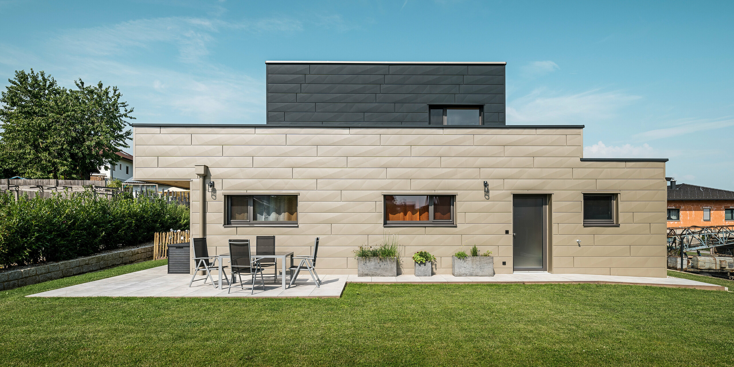 Gartenansicht eines modernen Einfamilienhauses in Wippenham, Österreich, mit einer zweifarbigen PREFA Aluminiumfassade. Die Fassade besteht aus Siding.X Fassadenpaneelen in den Farben Bronze und Anthrazit, die einen stilvollen Kontrast bilden und dem Gebäude eine moderne, klare Linienführung verleihen. Im Vordergrund ist eine großzügige Terrasse mit Gartenmöbeln zu sehen, die den gepflegten Rasen und das moderne architektonische Design des Hauses ergänzen.