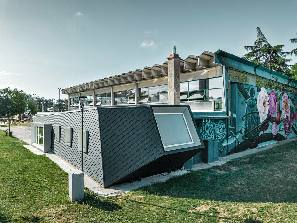 La palestra Rodriguez e la sua nuova estensione con il rivestimento in alluminio PREFA a forma di scaglie di pesce, vista di lato; il prato si estende davanti e di lato, mentre sullo sfondo si intravede in parte un campo sportivo.