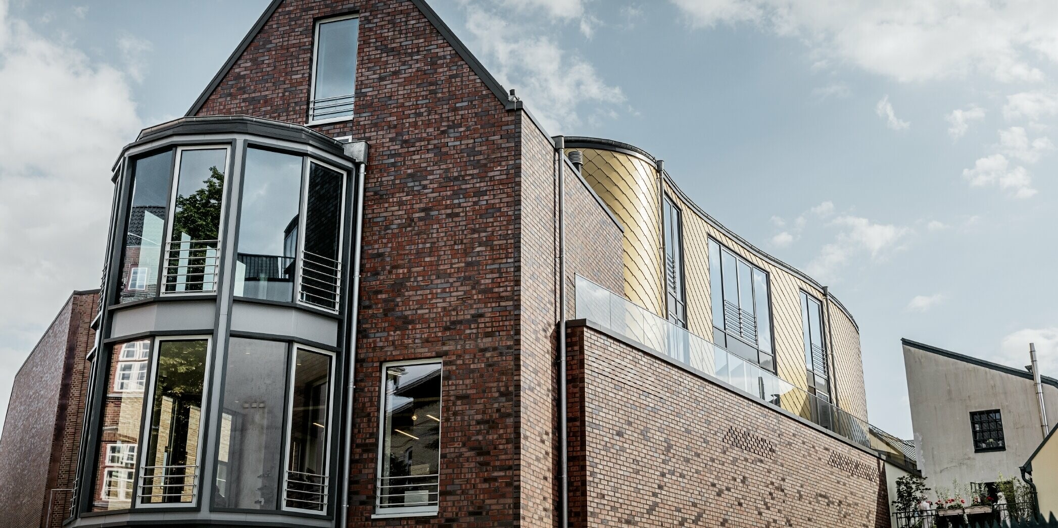 Geschäftshaus mit Backsteinfassade und großzügigen Glasflächen; Ein Teil der geschwungenen Fassade wurde mit der goldenen Wandraute aus Aluminium von PREFA verkleidet.