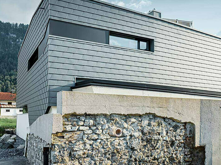 Fassadenverkleidung mit der PREFA Wandschindel aus Aluminium in Hellgrau mit einer Steinmauer vor dem Haus;