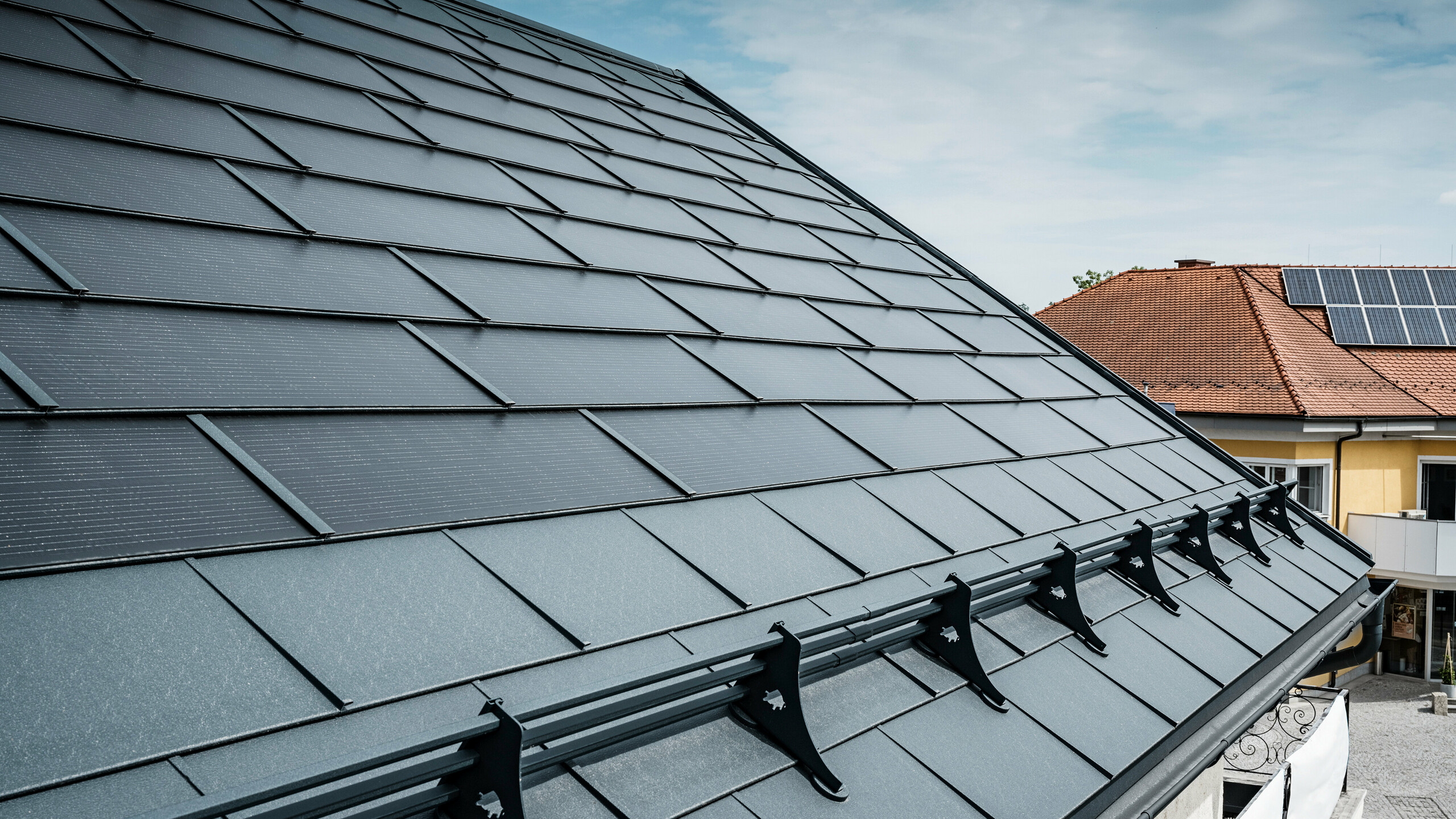 Vista di un tetto moderno con tegole fotovoltaiche PREFA color antracite. Vicino alla gronda, il tetto è stato dotato delle classiche tegole R.16 PREFA e di un sistema paraneve di PREFA. Sullo sfondo si vede il tetto di una casa vicina con tegole tradizionali e pannelli fotovoltaici sul tetto, che evidenzia il contrasto tra il design del tetto nuovo e quello tradizionale. L'innovativa tegola fotovoltaica PREFA combina estetica e funzionalità e offre una soluzione energetica sostenibile per tutti i tipi di edifici.