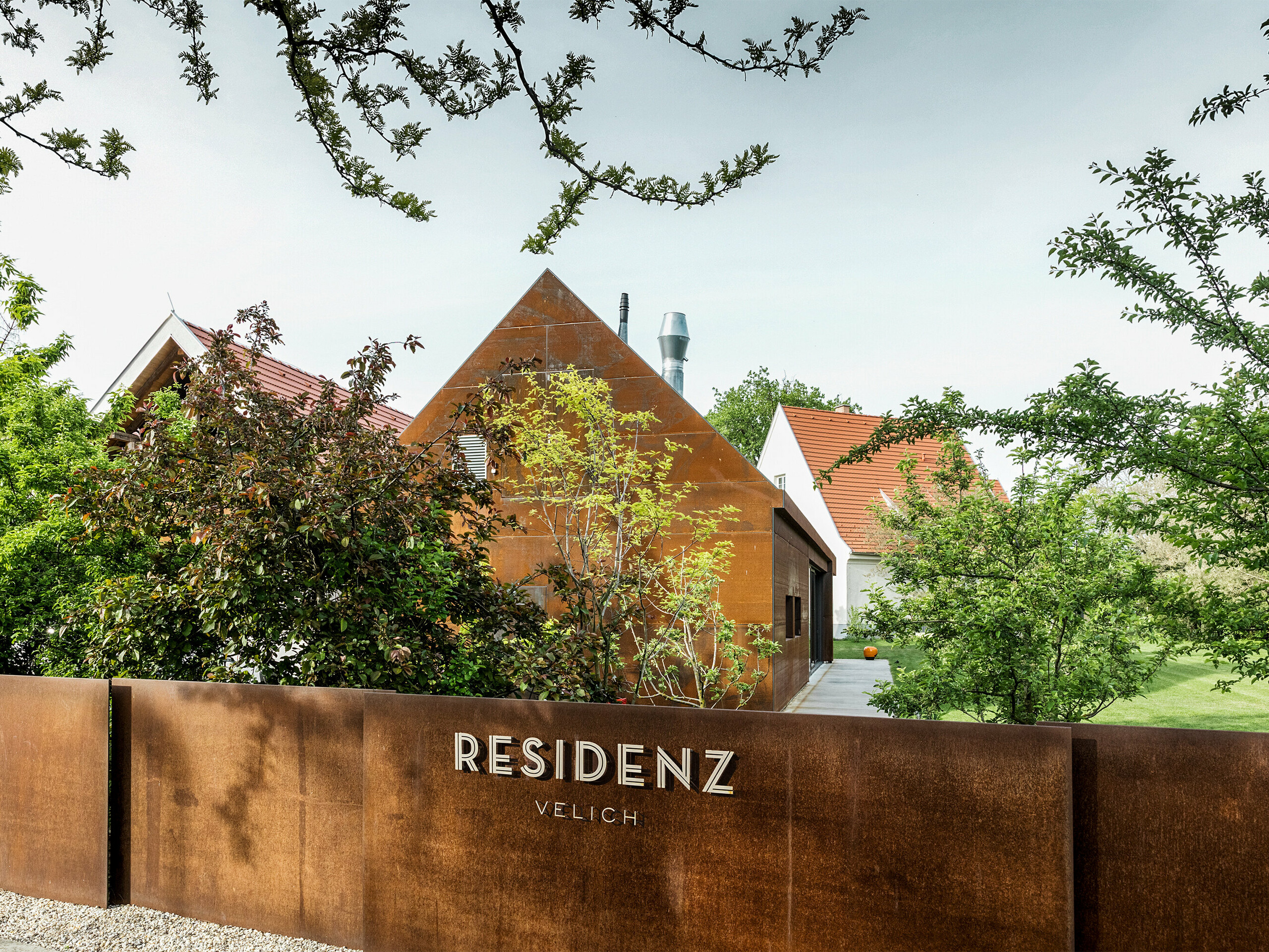 Residenza Velich nella regione del Seewinkel in Burgenland: Vista del moderno edificio, circondato da una rigogliosa vegetazione. La residenza funge da sede per il dialogo di esperti delle PREFARENZEN 2025, durante il quale architetti ed esperti di PREFA selezionano i più interessanti oggetti architettonici. In primo piano si vede una elegante parete metallica color ruggine con la scritta 'RESIDENZ VELICH'. L'architettura dell'edificio combina elementi tradizionali e moderni e si integra armoniosamente con l'ambiente naturale.