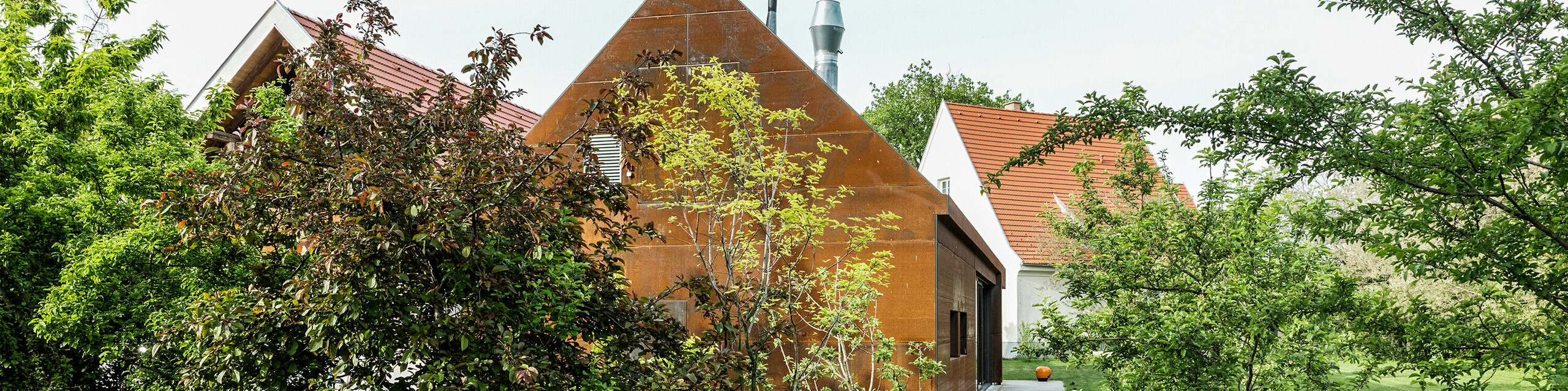 Rezidencia Velich v Seewinkel, Burgenland: pohľad na modernú budovu obklopenú bujnou vegetáciou. Rezidencia slúži ako miesto odborného dialógu PREFARENZEN 2025, v rámci ktorého architekti a odborníci z PREFA vyberajú najzaujímavejšie architektonické projekty. V popredí je vidieť štýlovú kovovú stenu hrdzavej farby s nápisom "RESIDENZ VELICH". Architektúra budovy spája tradičné a moderné prvky a harmonicky zapadá do okolitej prírody.