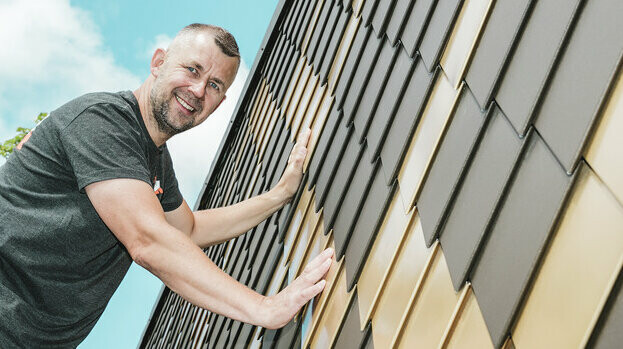 PREFA Handwerker mit grauem T-Shirt an einer PREFA Rauten-Fassade in Braun und Mayagold.