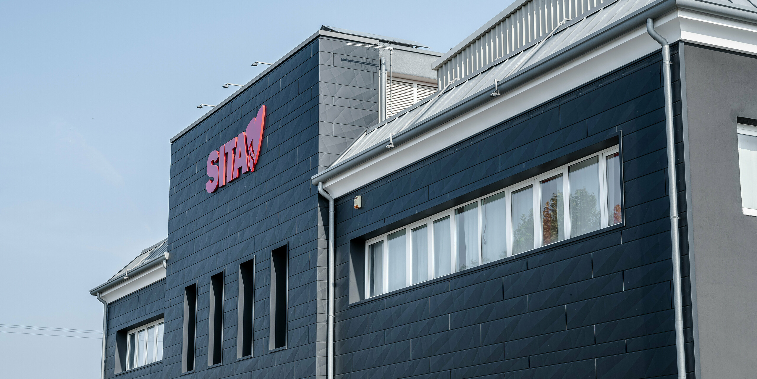 Schräge Ansicht des SITA-Firmengebäudes in Porcellengo, Italien, das mit der PREFA Siding.X Fassade in P.10 Schwarz verkleidet ist, welche die moderne Firmenarchitektur unterstreicht. Die Fassade kombiniert Funktionalität mit ästhetischem Design und bietet durch ihre einzigartige Oberflächenstruktur eine beeindruckende Optik. Das markant rote SITA-Logo hebt sich deutlich ab und verleiht dem Gebäude eine starke visuelle Identität. Die Aluminiumpaneele sind nicht nur ein Designelement, sondern auch ein Zeichen für nachhaltiges Bauen und Langlebigkeit.