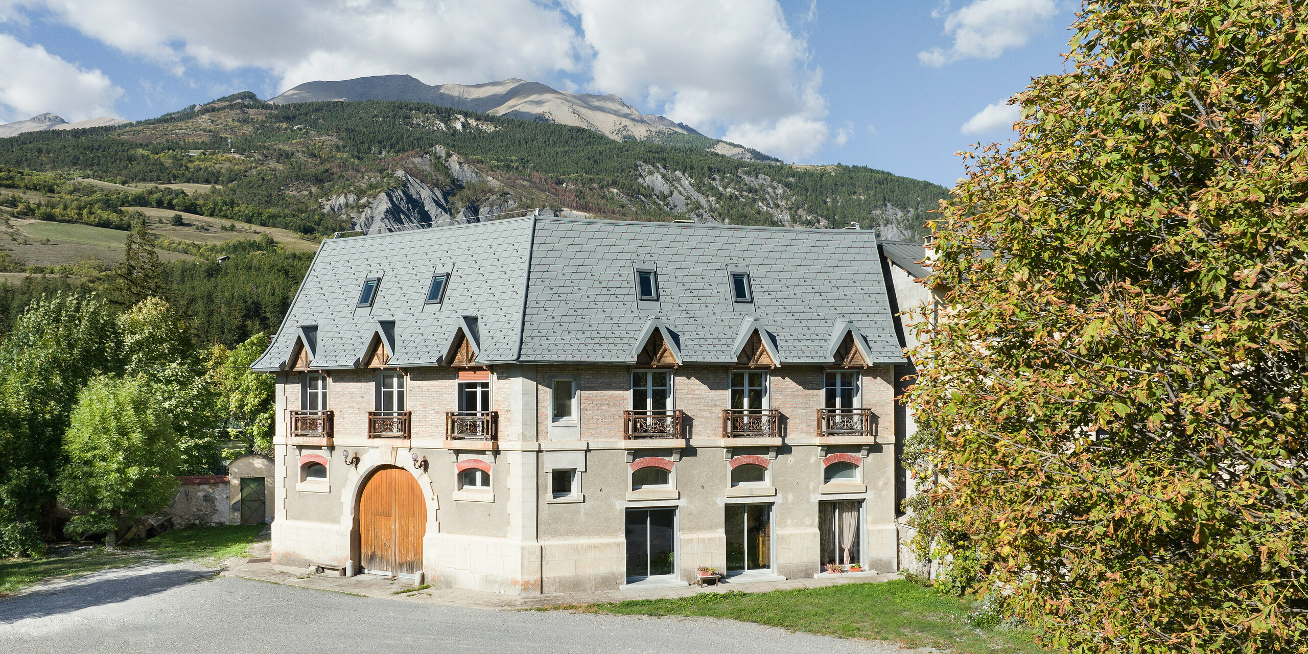 Frontansicht eines Anwesens in Barcelonnette, Frankreich. Das großflächige Dach wurde mit der PREFA Dachschindel in P.10 Hellgrau saniert. 