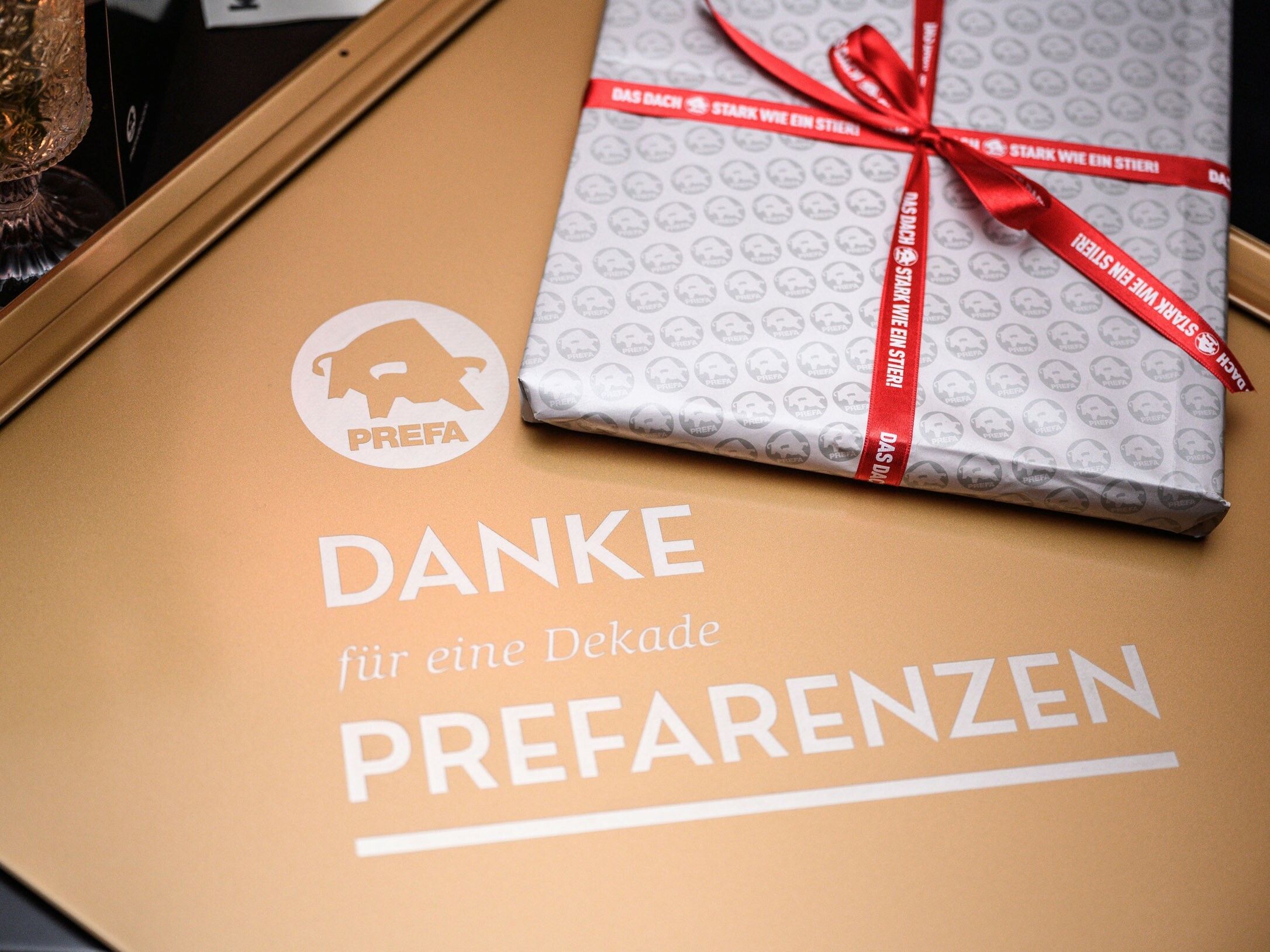 [Translate to Italiano:] Ein Geschenk liegt auf einer goldenen Oberfläche mit dem PREFA Logo und dem Schriftzug 'DANKE für eine Dekade PREFARENZEN'. Das Geschenk ist in silbernes Papier eingepackt, das mit kleinen PREFA Logos bedruckt ist, und mit einem roten Band gebunden, auf dem 'STARK WIE EIN STIER' steht