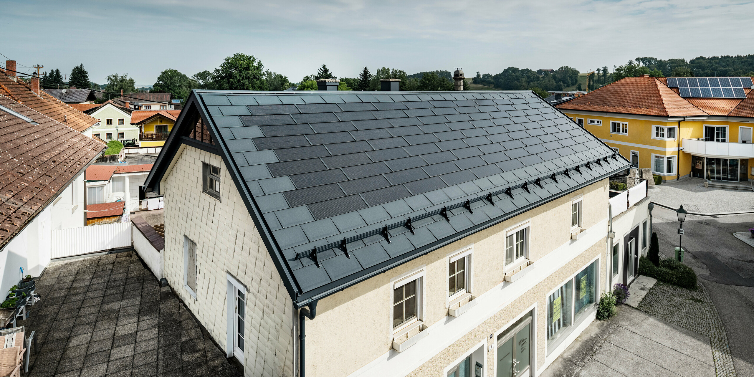 Vista dall'alto di una casa indipendente a Mettmach, in Austria. Il vecchio edificio è stato ristrutturato con tegole fotovoltaiche PREFA color antracite. I sistemi di protezione dalla neve e le linee chiare delle tegole fotovoltaiche sottolineano la lavorazione pulita dei prodotti in alluminio PREFA. Il tetto a tegole di colore rosso di un edificio vicino contrasta sullo sfondo. I moduli fotovoltaici sono installati sul tetto della vicina casa unifamiliare in una classica soluzione su tetto, a dimostrazione della diversità della progettazione di tetti ad alta efficienza energetica.