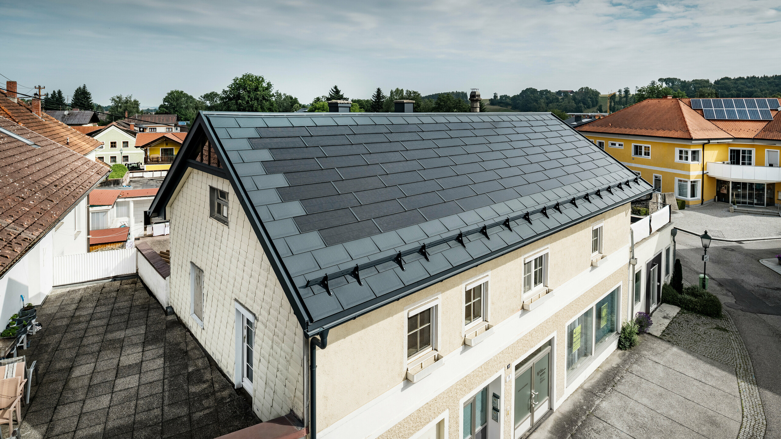 Vista dall'alto di una casa indipendente a Mettmach, in Austria. Il vecchio edificio è stato ristrutturato con tegole fotovoltaiche PREFA color antracite. I sistemi di protezione dalla neve e le linee chiare delle tegole fotovoltaiche sottolineano la lavorazione pulita dei prodotti in alluminio PREFA. Il tetto a tegole di colore rosso di un edificio vicino contrasta sullo sfondo. I moduli fotovoltaici sono installati sul tetto della vicina casa unifamiliare in una classica soluzione su tetto, a dimostrazione della diversità della progettazione di tetti ad alta efficienza energetica.