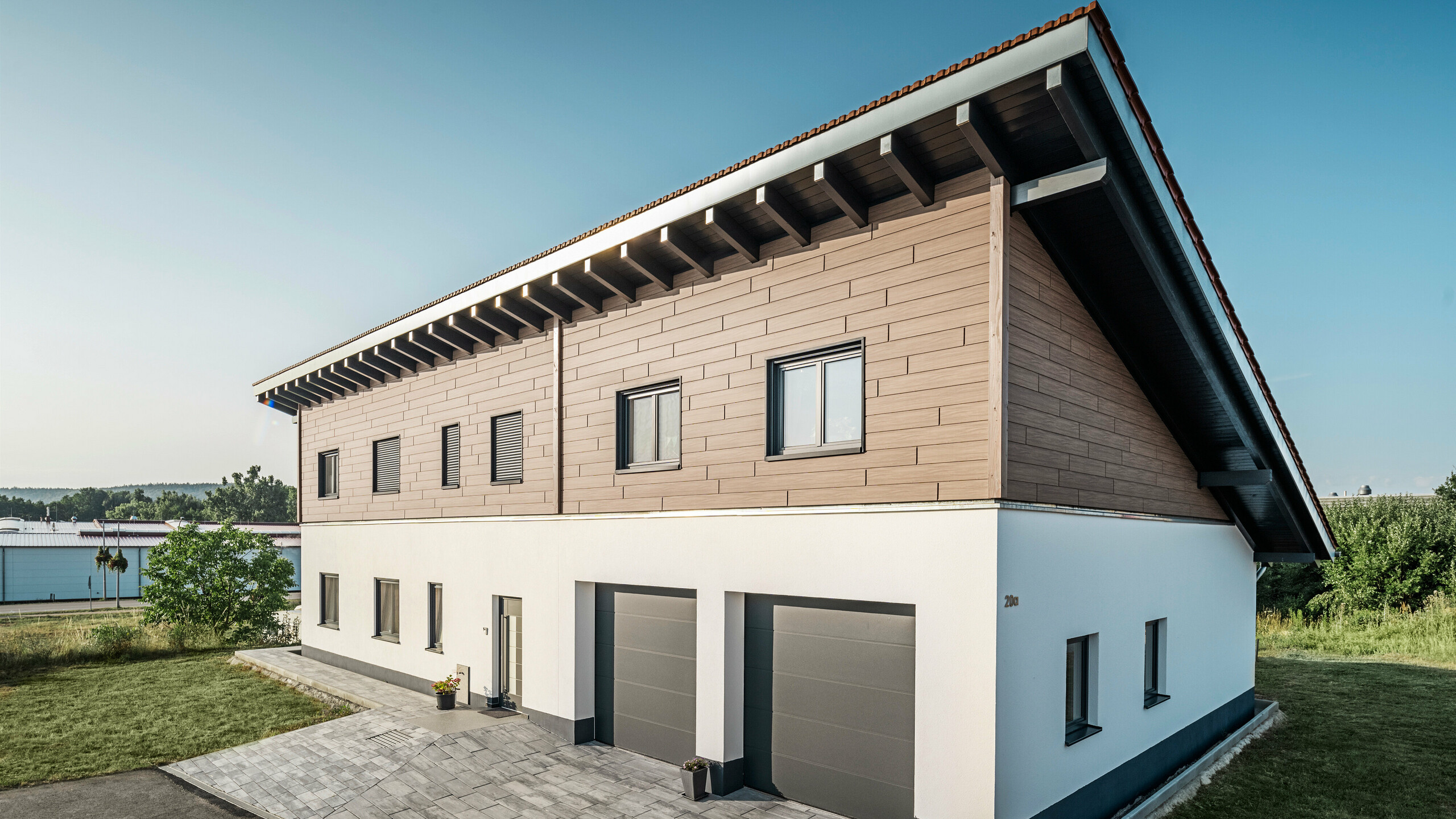 Vista laterale di una casa unifamiliare con tetto a due falde in Bruck. Il piano superiore è rivestito con PREFA doga in noce scuro. Sul lato destro dell'immagine sono visibili due porte di garage in colore antracite. La porta d'ingresso si trova al centro dell'edificio. Lo zoccolo è in tinta unita bianca. 