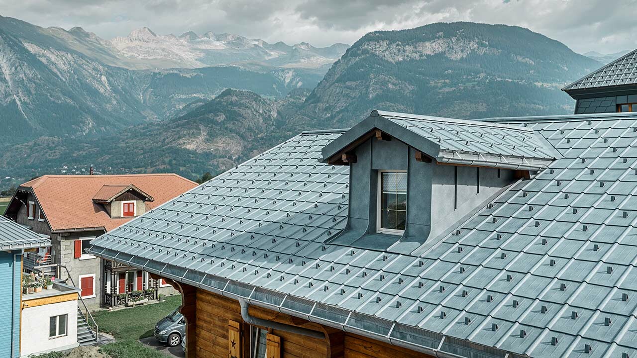 Tetto a sella con abbaino; il tetto in lamiera PREFA viene posato sulla superficie del tetto - la tegola in pietra grigia. L'abbaino è rivestito con aggraffatura PREFA in grigio pietra.