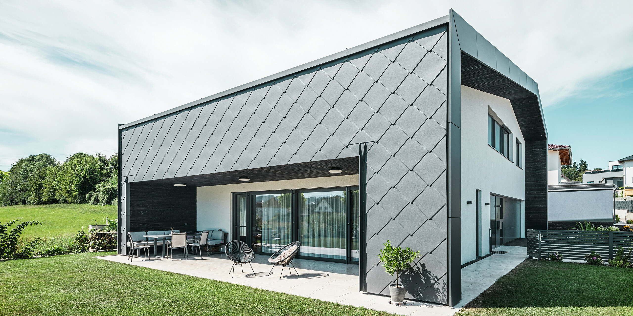 Gepflegter Gartenbereich eines Einfamilienhauses in Aschach. Das Gebäude ist stilvoll eingedeckt mit dunkelgrauen PREFA Aluminium-Wandrauten 44 × 44. Die überdachte Terrasse bietet einen gemütlichen Sitzplatz mit Blick auf die saftig grüne Rasenfläche. Die elegante Aluminiumverkleidung in Kombination mit weißen Putzflächen sorgt für eine moderne und witterungsbeständige Architektur. Große Fensterflächen lassen viel Tageslicht in den Innenraum und schaffen eine offene, lichtdurchflutete Atmosphäre.