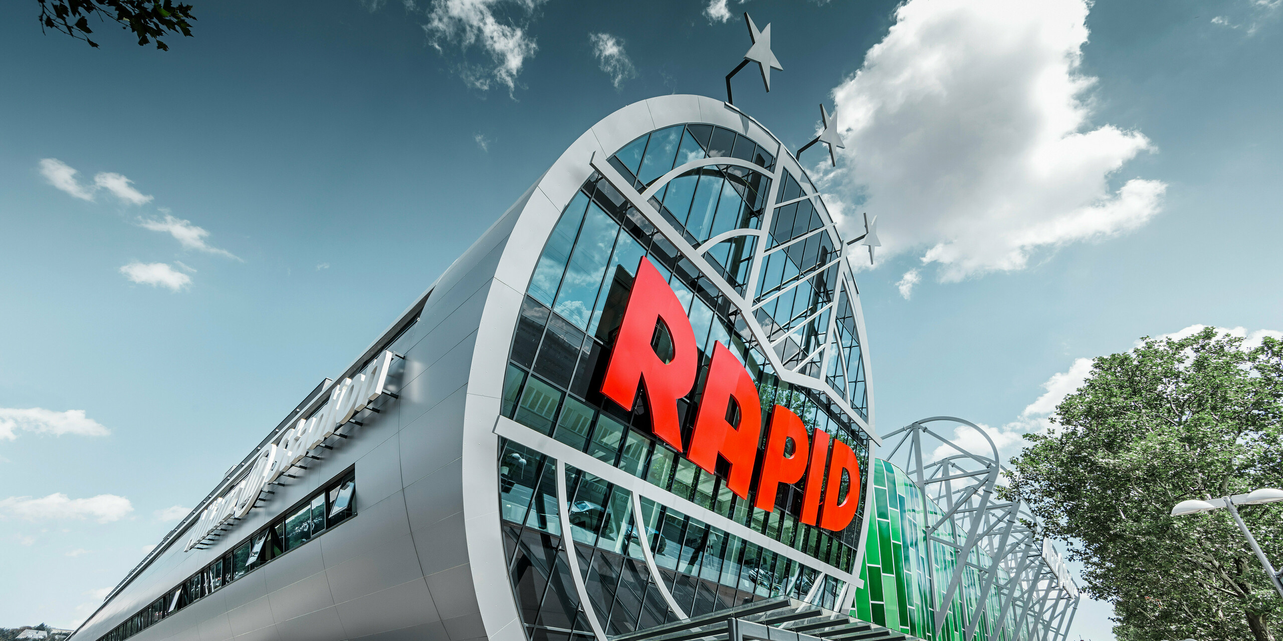 Ansicht des Allianz Stadions von SK Rapid Wien mit auffälliger PREFA Fassade in Form der Aluminium Verbundplatte in Silbermetallic. Die Gebäudehülle strahlt Dynamik und Moderne aus. Der Eingangsbereich, gekrönt mit dem leuchtend roten RAPID-Schriftzug, lädt ein in das Herz des Fußballs, eingefasst von einer futuristischen und zugleich eleganten Fassadenstruktur, die Sportarchitektur auf ein neues Niveau hebt.