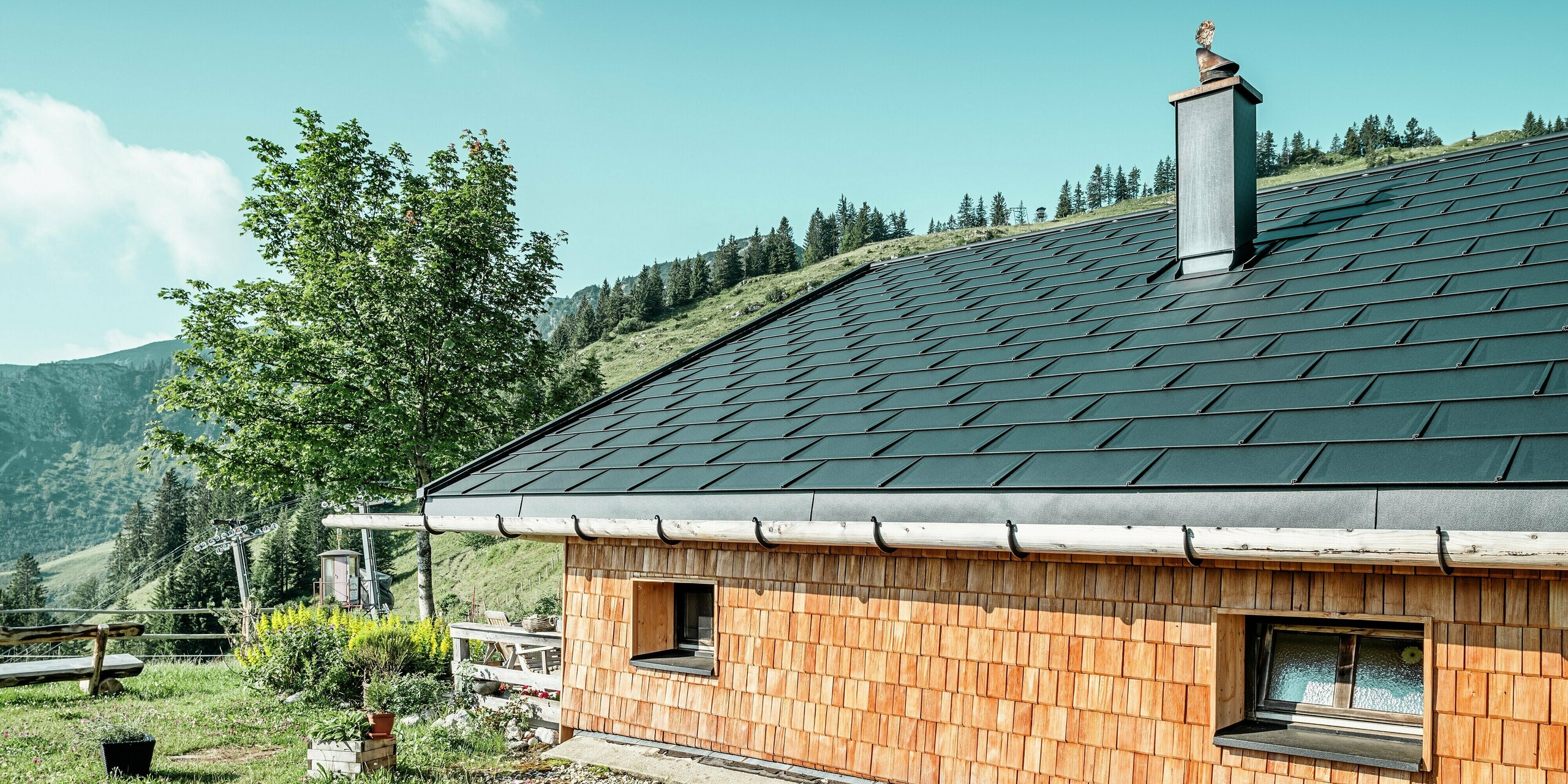 Seitenansicht der Birkehütte auf der Walleralm, Bayrischzell, nach der umfassenden Dachsanierung mit langlebigen PREFA Dachplatten R.16 in P.10 Schwarz und innovativen PREFA Solardachplatten. Der klassische Charme der Holzfassade wird durch die interessante Dachentwässerung mit einer Kombination aus Holzdachrinne und Aluminiumfallrohr abgerundet. Der elegante Kamin wurde mit Aluminium verkleidet und mit einer Metallfigur am höchsten Punkt versehen.Die kunstvolle Figur symbolisiert einen traditionellen Jagdhut. Die künstlerische Figur symbolisiert einen traditionellen Jagdhut. Die harmonische Verbindung von moderner Technik und traditioneller Architektur fügt sich perfekt in die malerische Berglandschaft ein.