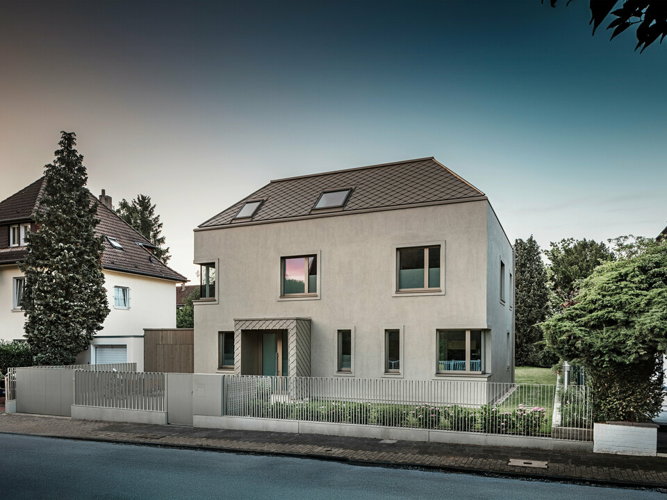 Una moderna casa indipendente con tetto a capanna e rivestimento di facciata e tetto in alluminio color bronzo di PREFA. La casa si trova in un tranquillo quartiere residenziale, circondato da giardini ben curati e da altre case unifamiliari. In primo piano si nota una bassa recinzione che separa la proprietà dalla strada. L'architettura della casa è caratterizzata da linee pulite e ampie finestre che lasciano entrare molta luce naturale all'interno. 