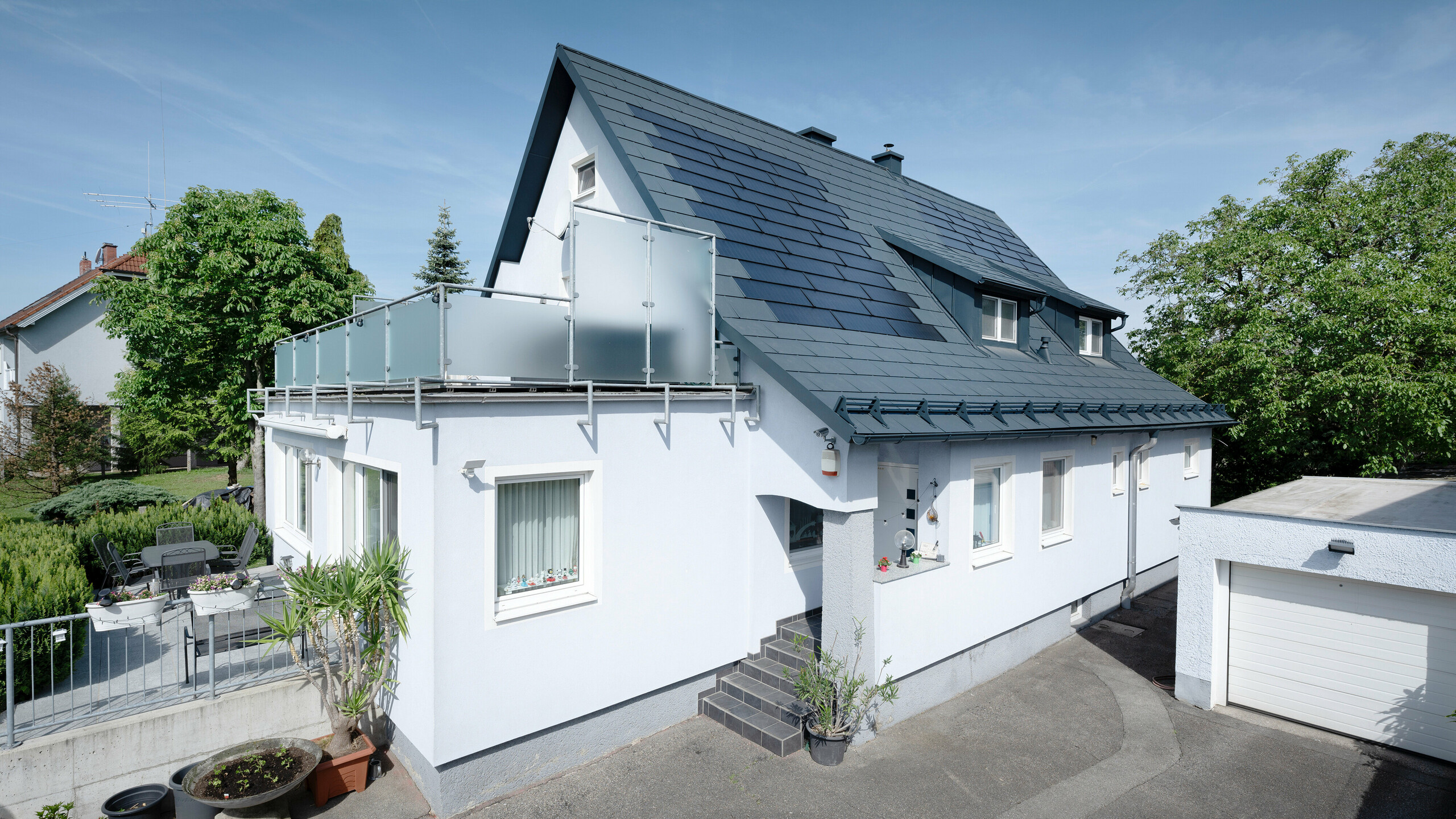 Dachsanierung eines Einfamilienhauses in Leonding mit PREFA Solardachplatten in Anthrazit. Das Bild zeigt das sanierte Haus aus einer seitlichen Perspektive mit klarer Sicht auf das Dach und die integrierten Solarmodule. Diese bieten eine nachhaltige und energieeffiziente Möglichkeit zur Stromerzeugung. Bei diesem Objekt erzeugen die Solardachplatten eine Stromleistung von 10,6 kWp. Die modernen, langlebigen und witterungsbeständigen PREFA Materialien tragen zu einer ästhetischen und funktionalen Dachkonstruktion bei, die den neuesten Standards für nachhaltiges Bauen entspricht.