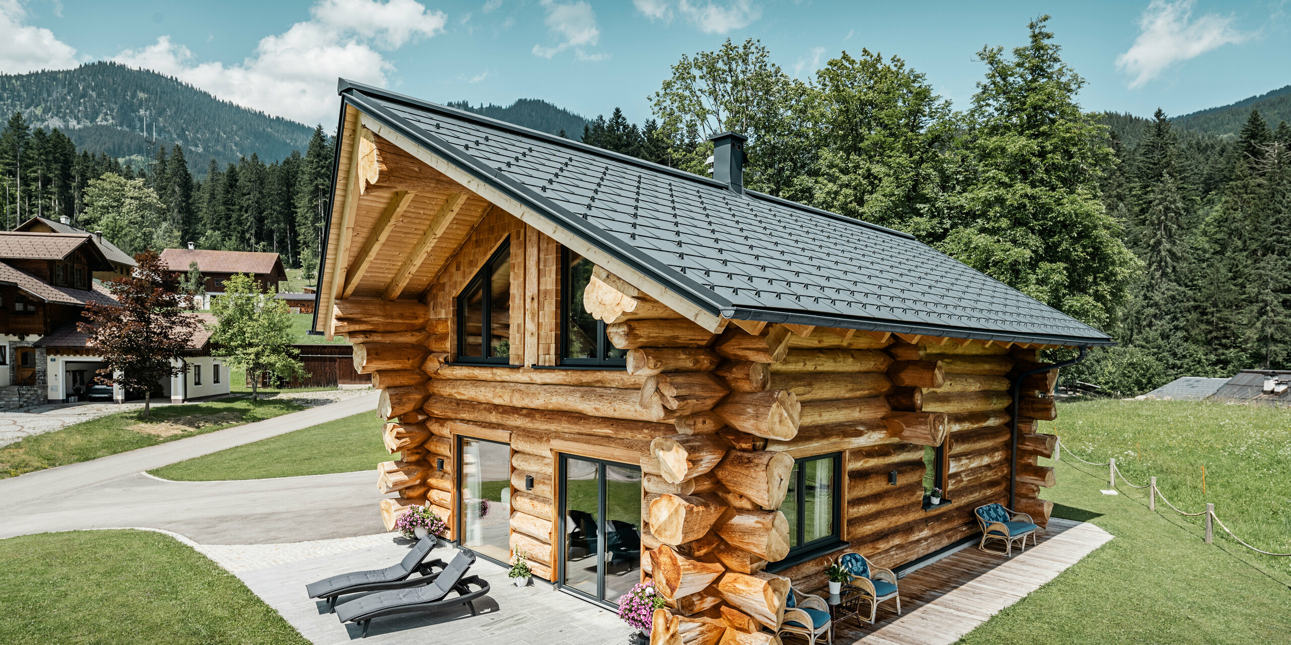 Rustikales Blockhaus in Gosau, Österreich, ausgestattet mit langlebigen PREFA Dachplatten R.16 in P.10 Anthrazit. Die kleinformatigen Dachplatten aus Aluminium sind harmonisch eingebettet in die natürliche Landschaft mit üppigen grünen Bäumen und malerischen Bergen im Hintergrund. Das hochwertige, anthrazitfarbene Dachsystem bietet einen starken Kontrast zu den traditionellen Holzblockwänden des Hauses und ist optimiert für Witterungsbeständigkeit und Langlebigkeit. Dieses Bild veranschaulicht die perfekte Kombination aus traditioneller Bauweise und modernem Design von PREFA.