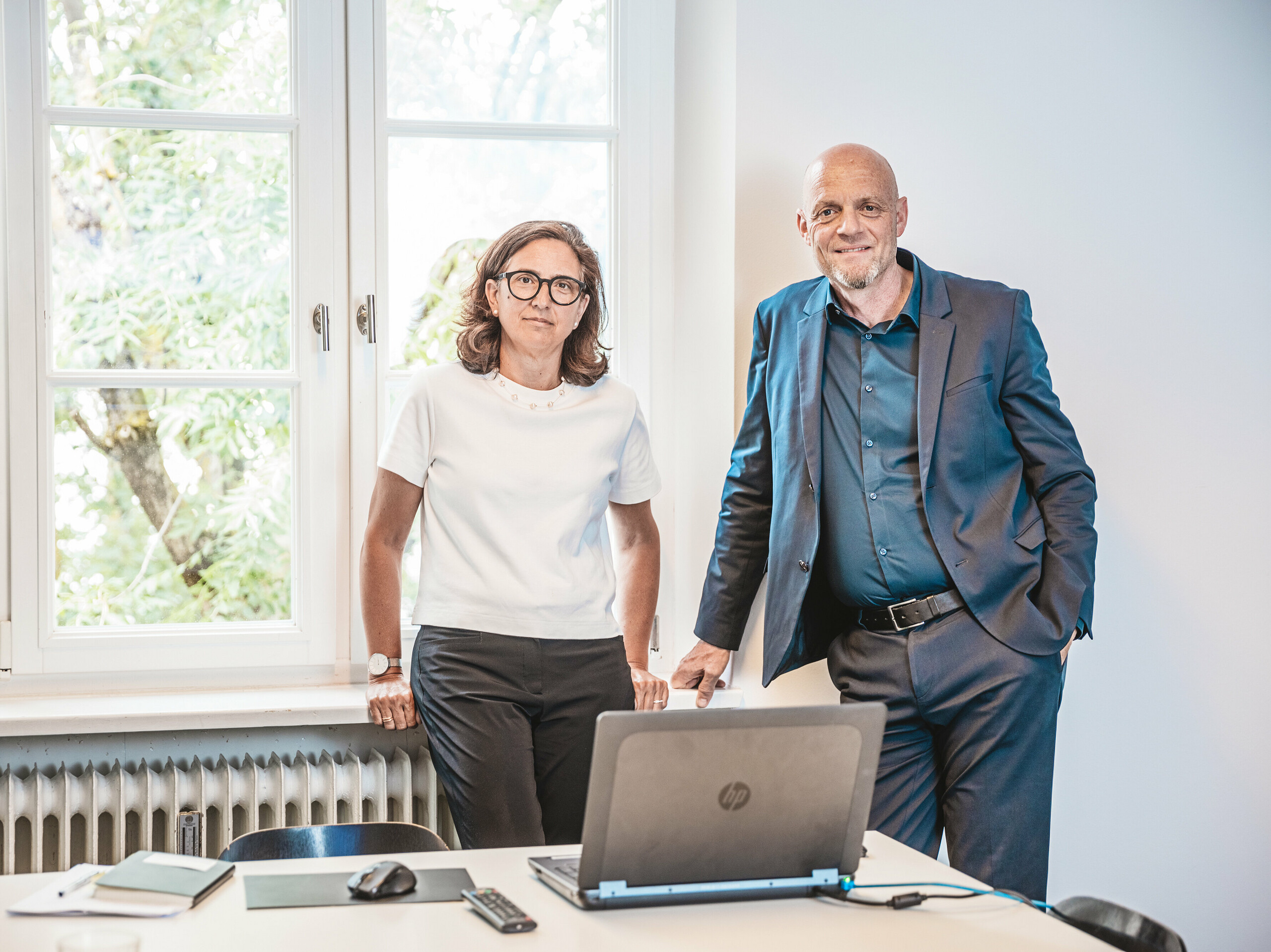 Patricia Ezcura e Gerold Heugenhauser dello studio WWA Architekten di Monaco, le menti creative del progetto residenziale di Utting am Ammersee. I due sono in piedi in un ufficio, dietro un tavolo con un computer portatile e dei quaderni e davanti a una grande finestra. La loro pianificazione e progettazione innovativa del moderno complesso edilizio sul sito di Schmucker, con l'impiego di prodotti sostenibili PREFA come le tegole R.16, è caratterizzata da un'integrazione armoniosa nell'ambiente circostante e da un'architettura esteticamente gradevole.