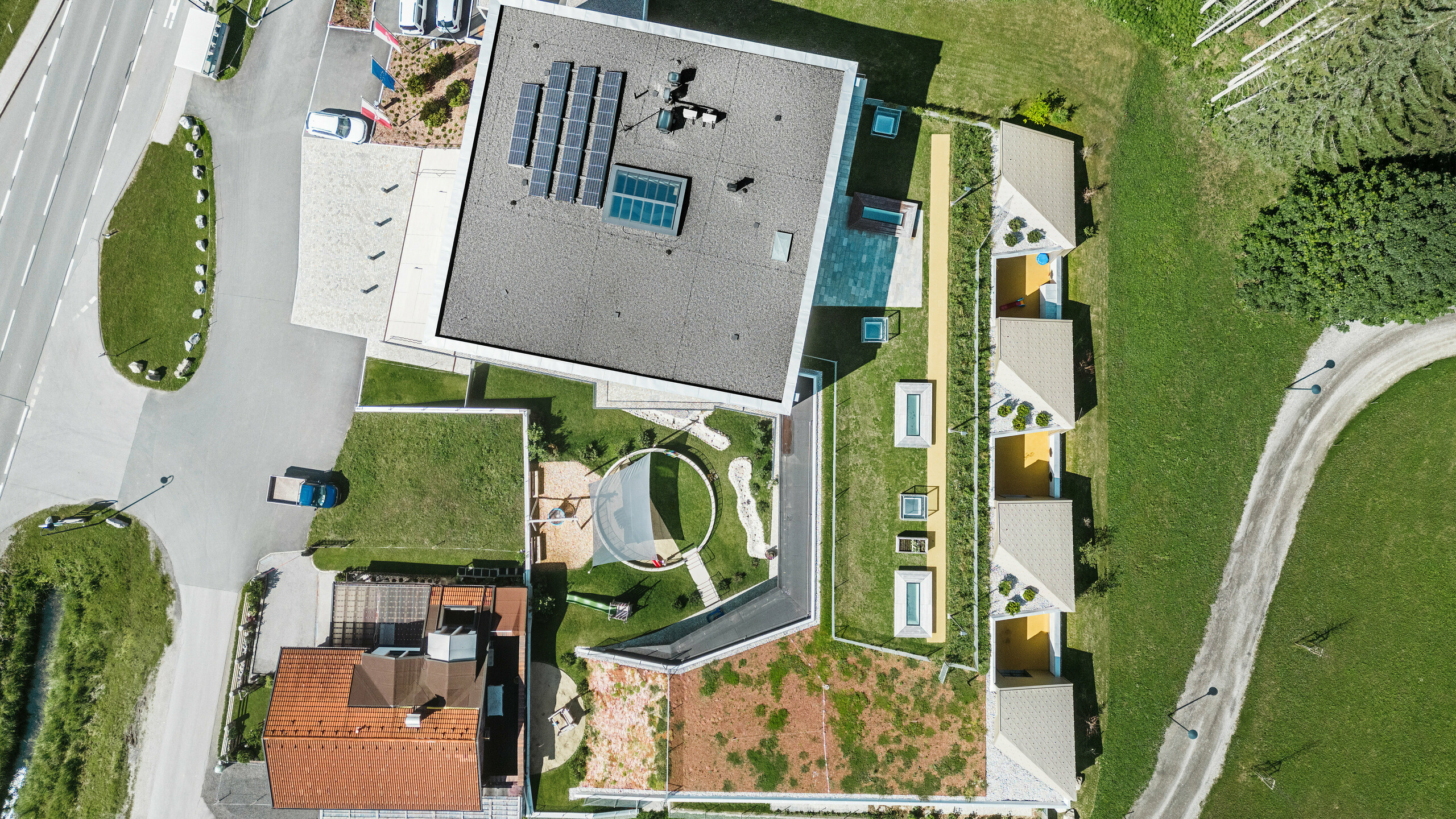 Vogelperspektive auf das Bildungszentrum von Gerlos mit einem Ensemble aus modernen und traditionellen Gebäuden. Das Hauptgebäude präsentiert sich mit einem grauen, mit Solarpaneelen bestückten Schrägdach und ist von weiteren Gebäuden mit gelben Fassaden und grünen Dächern umgeben. Im Hintergrund zeichnet sich die malerische, waldreiche Alpenlandschaft ab, während sich im Vordergrund geparkte Autos und eine gewundene Straße befinden.