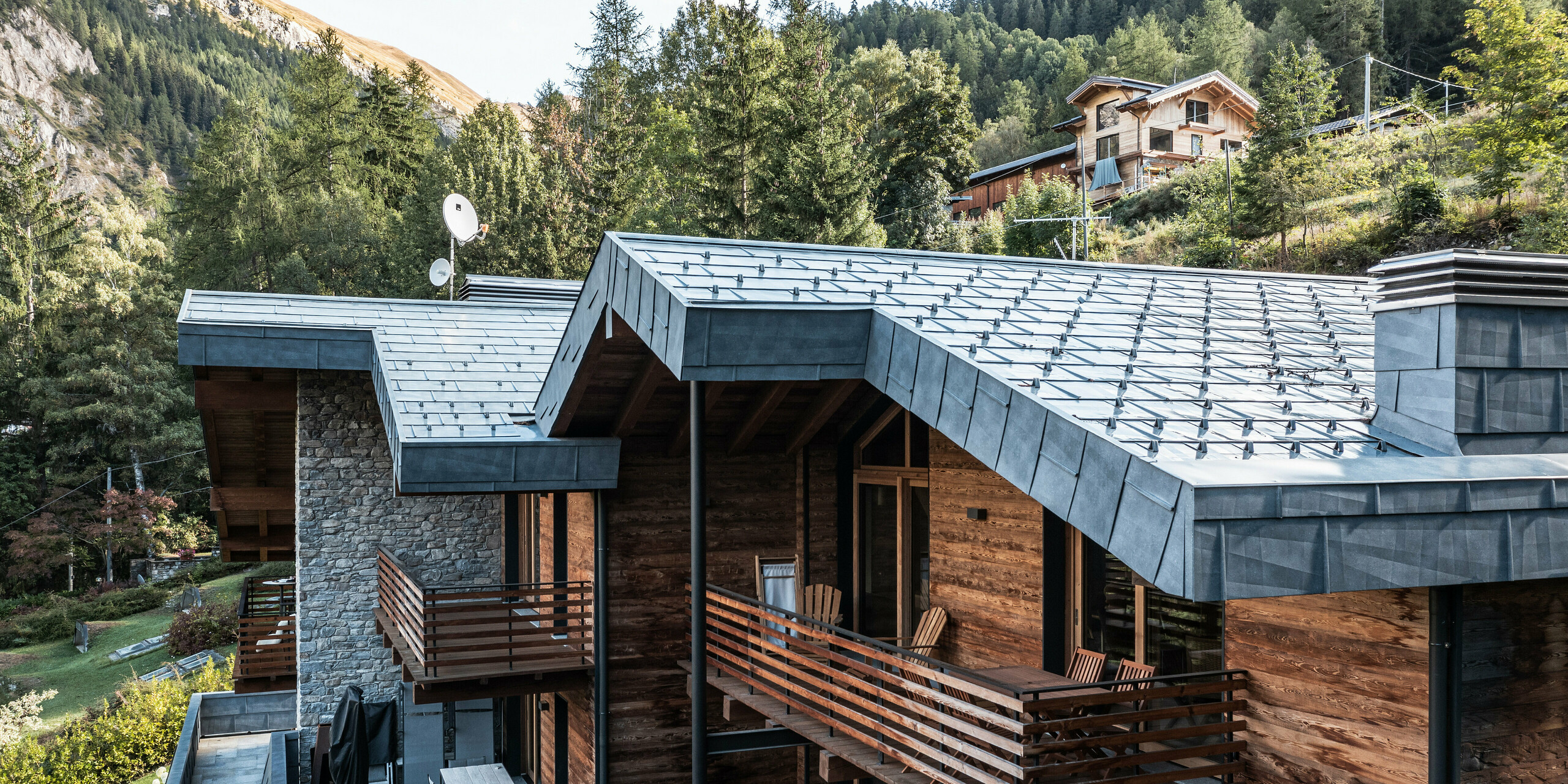Komplexní vila v Courmayeur s dřevěnými fasádami a hliníkovými střešními panely PREFA FX.12 v barvě P.10 břidlicové. Architektura kombinuje moderní a tradiční prvky, jako je zdivo z přírodního kamene a vícepatrové dřevěné konstrukce, doplněné stylovými hliníkovými detaily. Několik balkonů a teras poskytuje dostatek venkovního prostoru doplněného svěží zelení a dramatickou horskou krajinou za nimi.