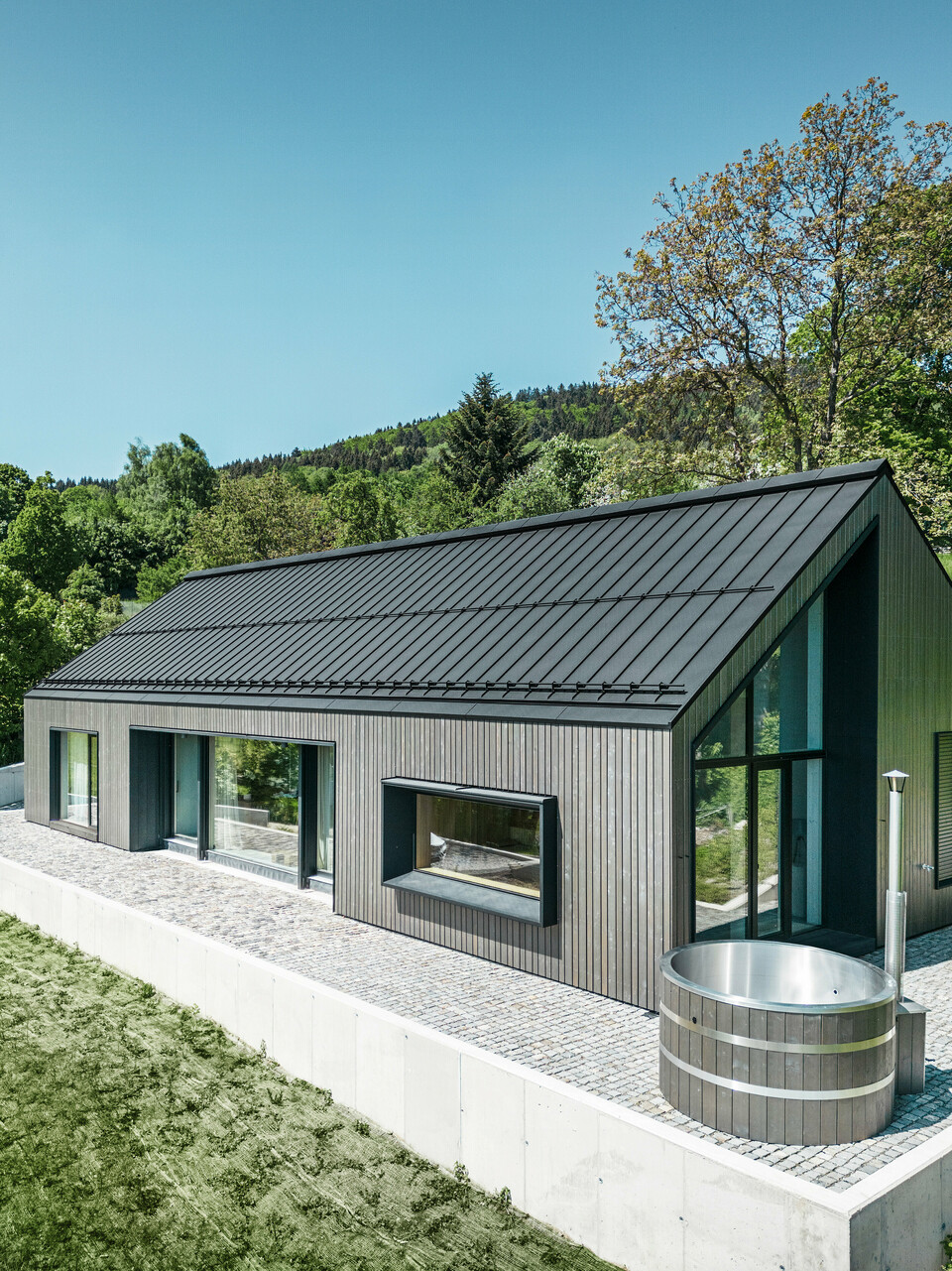 Ein modernes Holzmassivhaus in Kuncice (CZ) wurde mit großen Fensterflächen ausgestattet. Das dunkle Holzmassivhaus verfügt über ein Dachsystem aus PREFALZ in der Farbe P. 10 Schwarz. Im hinteren Außenbereich steht ein Whirlpool mit einer Holzverkleidung.