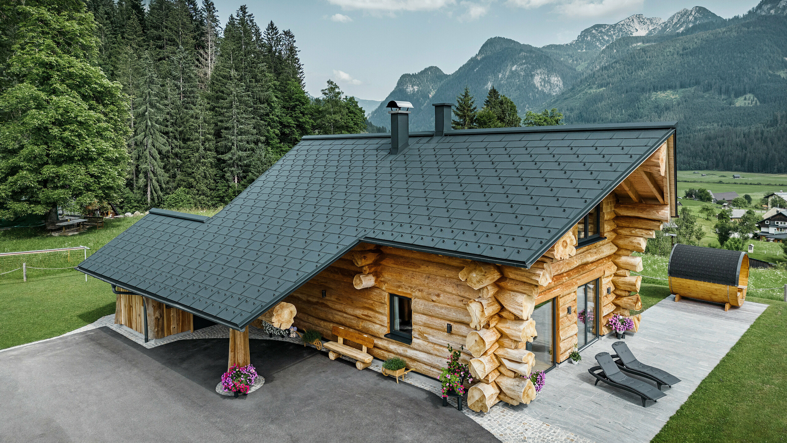 Schräge Ansicht eines malerischen Blockhauses in Gosau, Österreich, ausgestattet mit einem PREFA Dach aus anthrazitfarbenen Dachplatten R.16. Die Architektur kombiniert rustikales Blockhausflair mit modernen Elementen, umgeben von dichten grünen Wäldern und einer beeindruckenden Bergkulisse. Das Haus strahlt mit seiner ländlichen Eleganz und den präzise verlegten Dachelementen aus Aluminium, die für ihre Langlebigkeit und Witterungsbeständigkeit bekannt sind, eine einladende Atmosphäre aus.