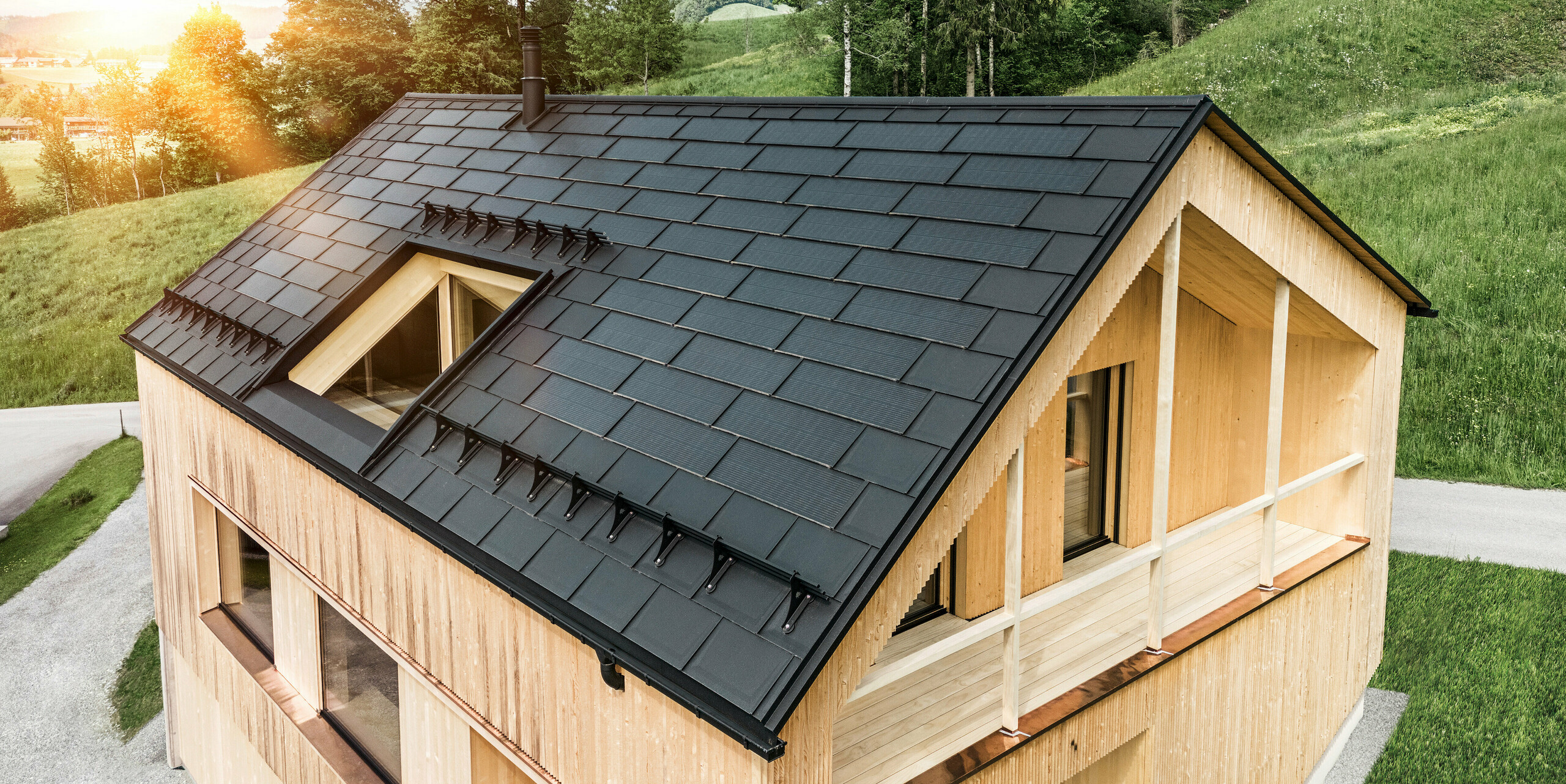 Vista di una moderna casa sostenibile in legno a Egg con un tetto PREFA Solar che si distingue per il suo aspetto scuro, strutturato ed elegante. Le tegole fotovoltaiche sono posizionati strategicamente trale classiche tegole del tetto R.16 per massimizzare il guadagno solare e allo stesso tempo armonizzarsi esteticamente con il design complessivo. La casa è in armonia con il verde circostante, mentre il caldo sole serale enfatizza la texture del legno naturale della facciata e sottolinea la solidità della costruzione. Un esempio armonioso di costruzione attenta all'ambiente e all'efficienza energetica.