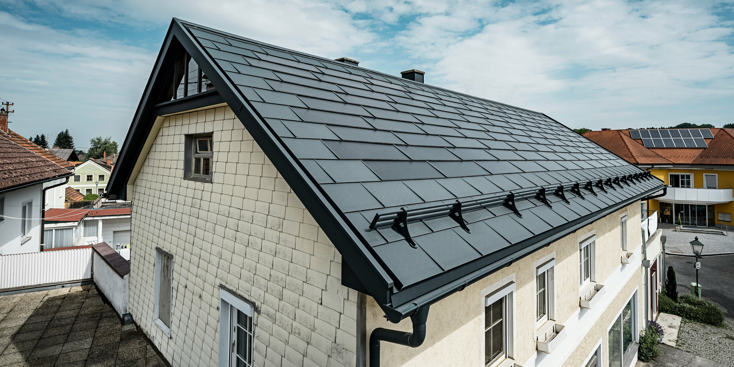 Vista dall'alto del tetto di una casa indipendente a Mettmach, in Austria. La casa è stata dotata di tegole fotovoltaiche PREFA di colore antracite nell'ambito di un progetto di ristrutturazione. La superficie strutturata delle tegole si armonizza con la semplice eleganza della casa. Sul bordo del tetto è stato applicato un sistema a tubi fermaneve. Sullo sfondo sono visibili altre case con soluzioni di tetti tradizionali e solari, che sottolineano la progettazione di tetti moderni ed efficienti dal punto di vista energetico in un ambiente rurale.