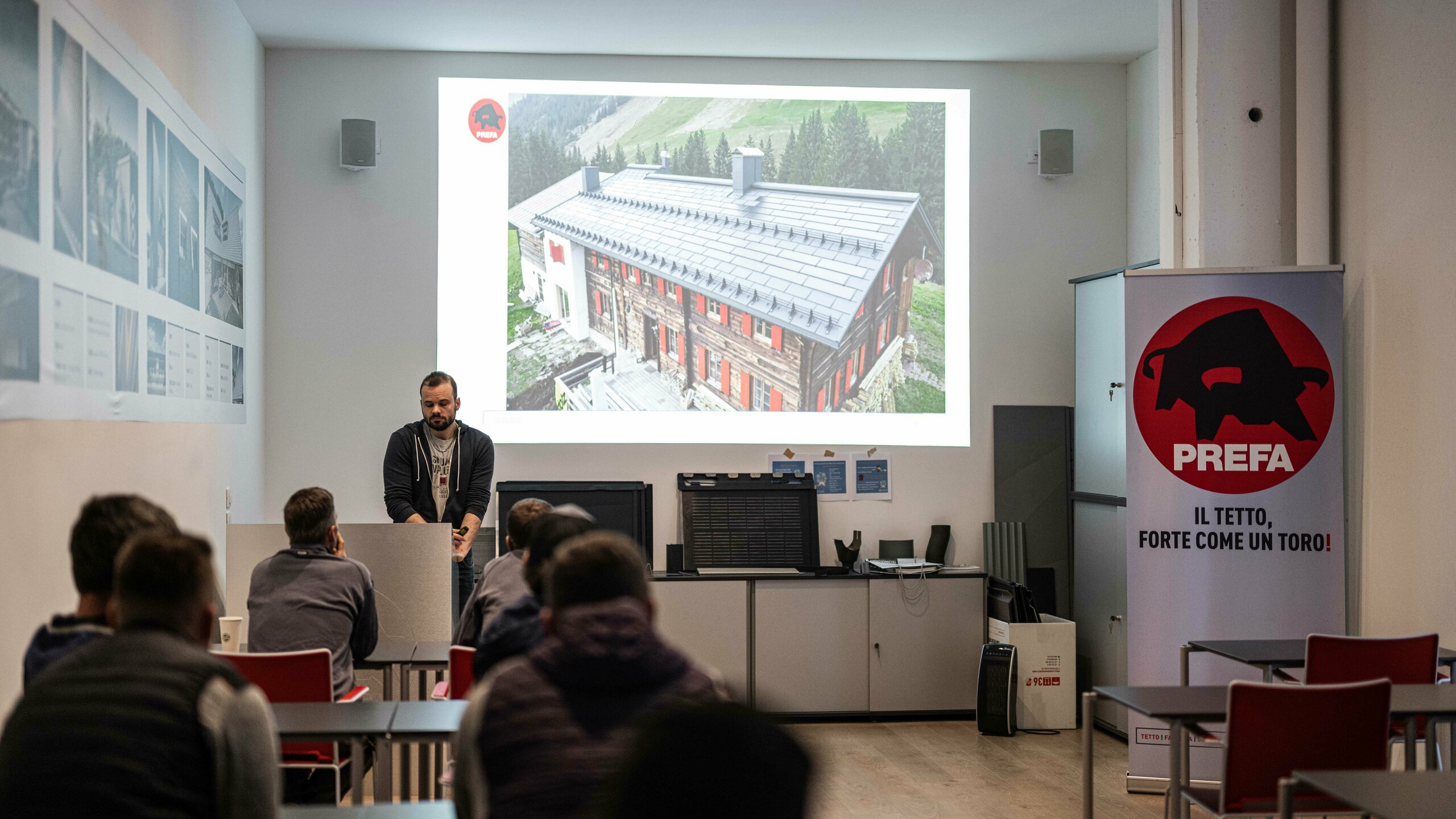 Un docente dell'Accademia PREFA presenta le soluzioni PREFA per tetti e facciate. Sullo schermo si vede l'immagine di una casa con i tegola fotovoltaica PREFA. I partecipanti siedono nella sala di formazione e seguono la presentazione. Nella sala c'è uno striscione PREFA con lo slogan “Il tetto, forte come un toro!”. (Il tetto, forte come un toro!) è visibile.