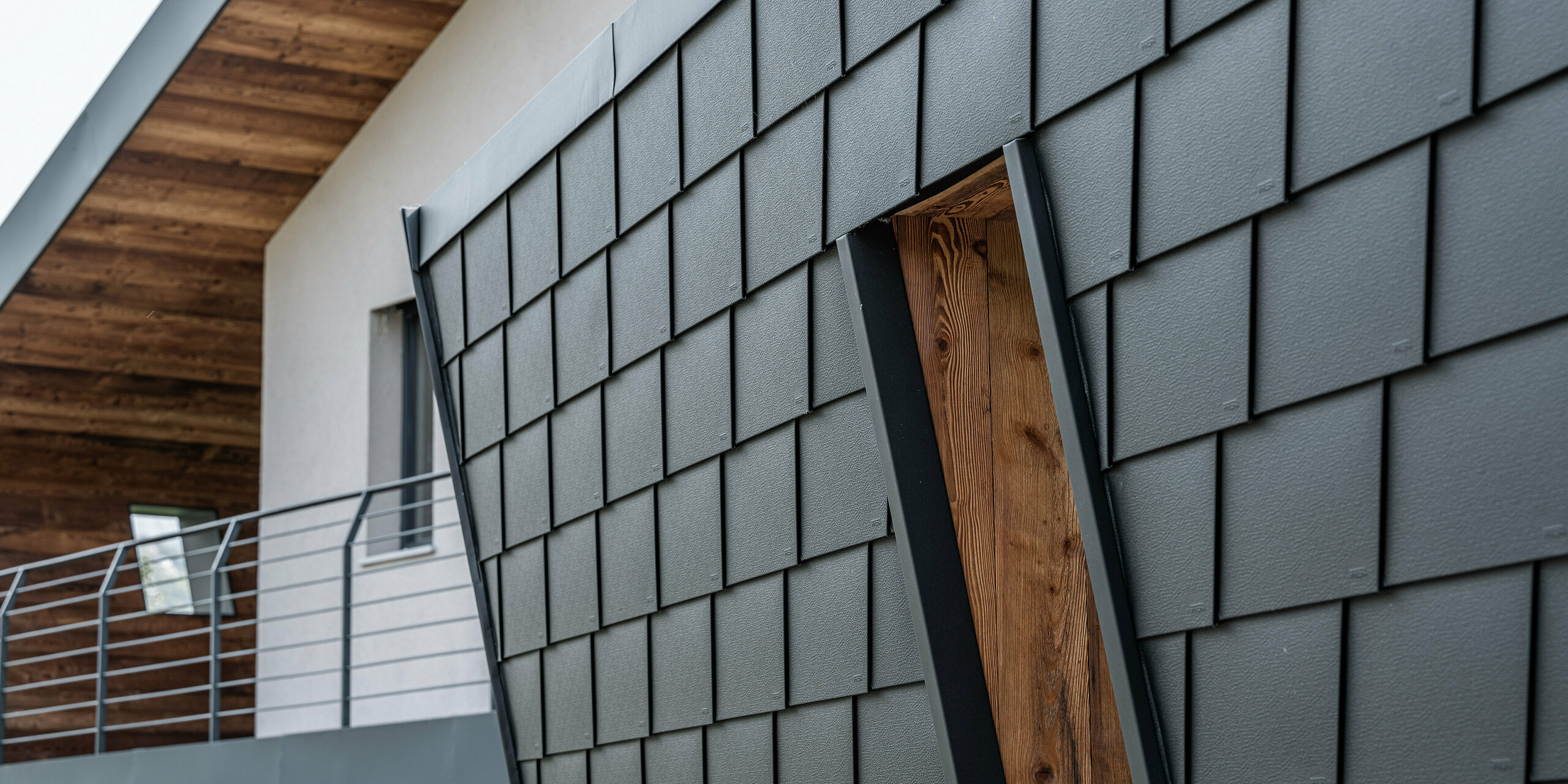 Detailaufnahme der Villa Tia in Italien mit einem PREFA Fassadensystem. Die klassischen Dach- und Wandschindeln von PREFA sind in einem versetzten Muster angeordnet, das durch die sorgfältige Verarbeitung und die dunkle Farbgebung eine moderne Textur erzeugt. Der Kontrast zwischen den Aluminiumpaneelen und den warmen Holzelementen des Dachüberstands vermittelt eine harmonische Verbindung von traditionellen und zeitgenössischen Baustoffen.