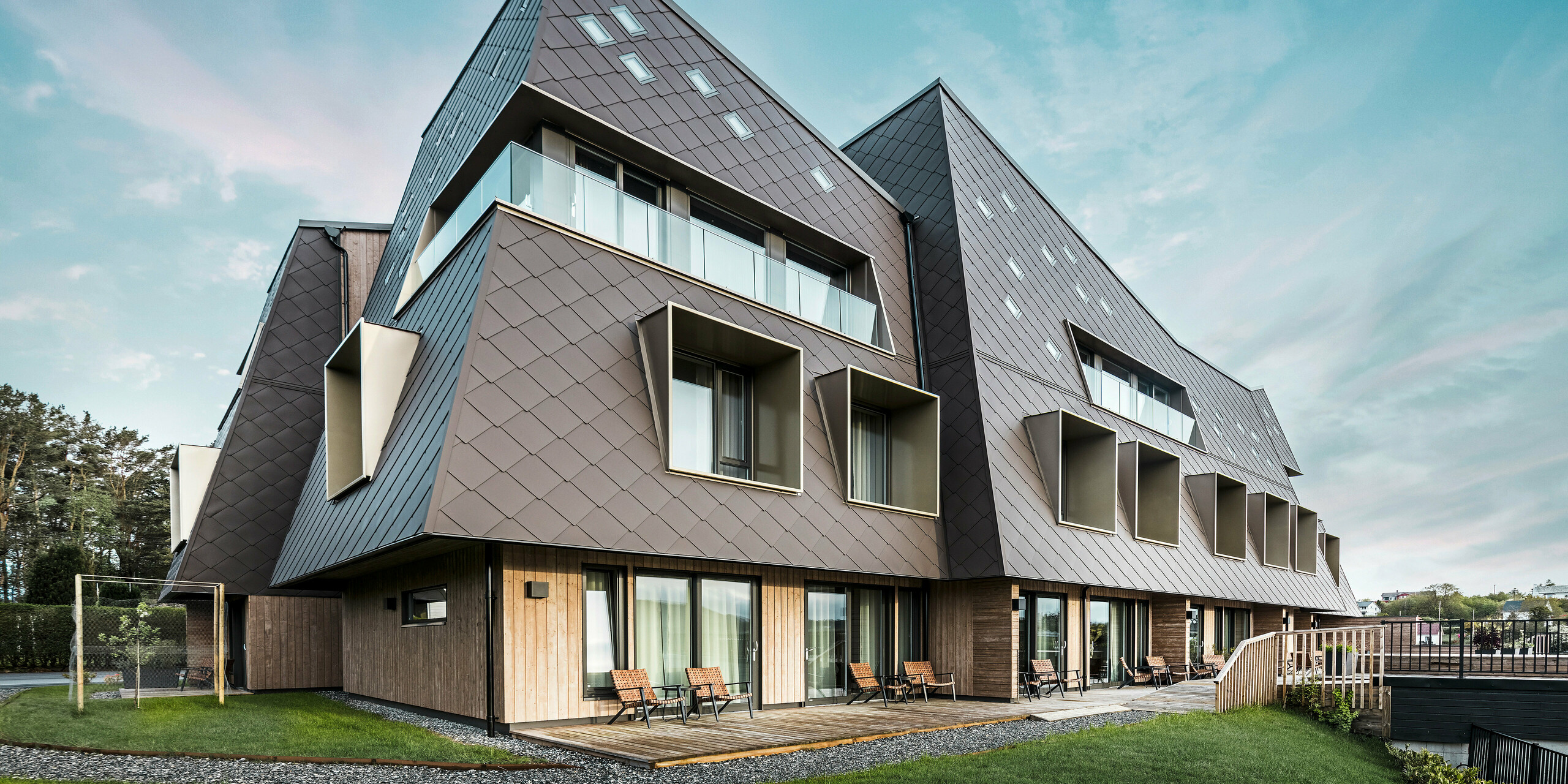 Froschperspektive des Boutiquehotels Beckerwyc House in Bekkjarvik, Norwegen, mit einer markanten Fassade aus PREFA Wandrauten 44 × 44 in Nussbraun. Die außergewöhnliche Architektur des Gebäudes, mit schrägen Fassadenelementen und hervortretenden Fenstern, schafft ein futuristisches Erscheinungsbild, das sich harmonisch in die natürliche Umgebung einfügt. Die moderne Gestaltung und die Verwendung hochwertiger Materialien betonen das exklusive Design des Hotels.