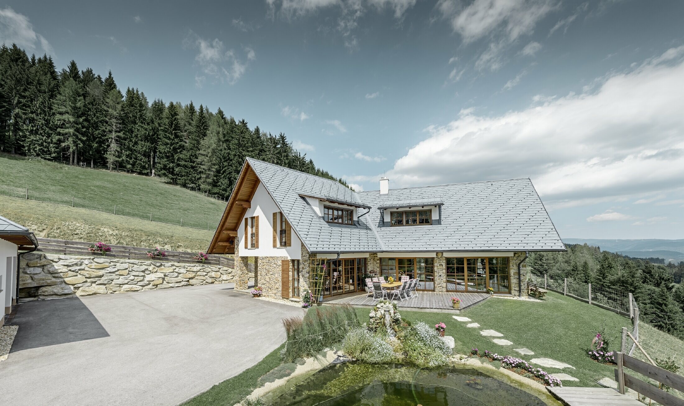 La casa unifamiliare in pendenza, costruita a forma di L, ha un tetto a due falde con due abbaini. Il tetto è stato rivestito con la scandola PREFA in grigio pietra. In primo piano c'è un laghetto e sullo sfondo c'è un bosco.