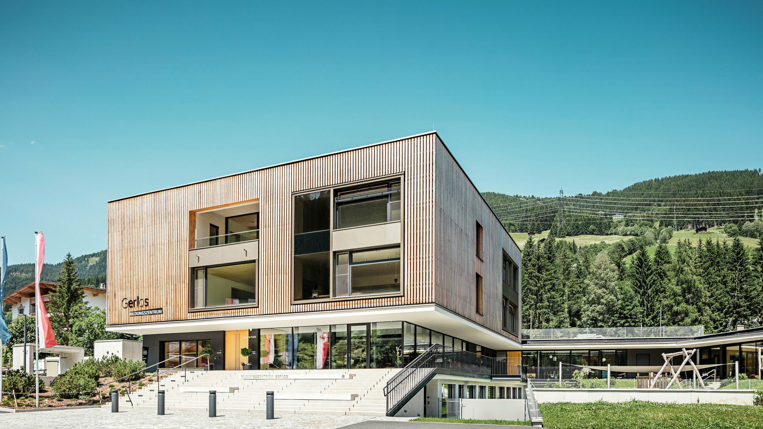 Frontansicht des modernen Bildungszentrum mit Holzfassade.  Das Gebäude ist zweistöckig mit großen Fensterfronten und einem flachen Dach, das sich in die umliegende Berglandschaft mit dicht bewaldeten Hügeln nahtlos einfügt. Im Vordergrund sind gepflegte Grünflächen, eine gestufte Zufahrt und Werbefahnen zu sehen. Das Design vermittelt eine Verbindung von Funktionalität und ästhetischer Architektur in harmonischem Einklang mit der Natur.