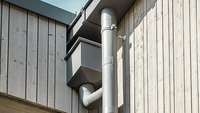 Primo piano del sistema di smaltimento per tetti PREFA in P.10 grigio scuro su un edificio residenziale nel sito di Schmucker a Utting am Ammersee. Il sistema di raccolta dell'acqua, moderno e funzionale, si integra perfettamente con la facciata in legno dell'edificio, sottolineando la lavorazione di alta qualità e il design sofisticato. L'immagine si concentra sulla robusta cassetta di raccolta dell'acqua e sui tubi di scarico, elegantemente collegati tra loro. I materiali durevoli e l'installazione precisa dei prodotti in alluminio PREFA garantiscono un deflusso efficace e contribuiscono all'architettura sostenibile dell'edificio ibrido in legno.