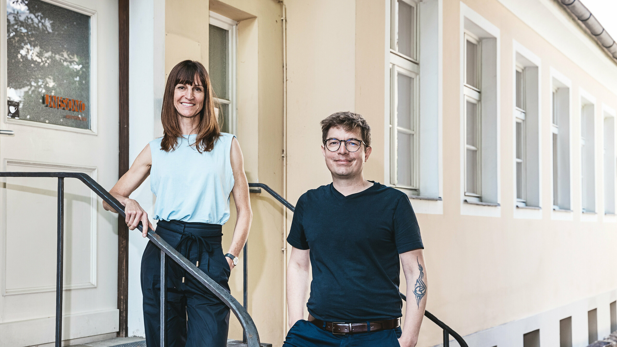 I principali architetti dello studio UNISONO ARCHITEKTEN di Innsbruck sono in piedi su una scala di pietra di fronte a un edificio dipinto di beige con finestre bianche. La donna indossa una camicetta blu senza maniche e pantaloni neri, l'uomo una maglietta nera e pantaloni blu con cintura marrone. Sorridono amichevolmente all'obiettivo, il che indica un'atmosfera aperta e accogliente.