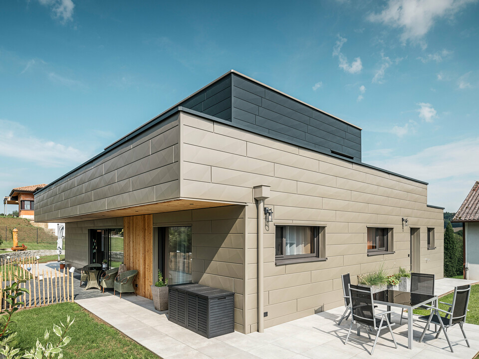 Ein modernes Einfamilienhaus in Wippenham, Österreich, mit einer zweifarbigen PREFA Aluminiumfassade, die aus den extravaganten Siding.X Fassadenpaneelen in Bronze und Anthrazit besteht. Die Fassade zeigt klar erkennbare, elegante Linien und Kantungen. Deutlich sichtbar ist die harmonisch integrierte PREFA Dachentwässerung mit Wasserfangkasten und Ablaufrohr, die sich nahtlos in das architektonische Gesamtkonzept einfügt. Im Vordergrund ist eine gemütliche Terrasse mit Gartenmöbeln zu sehen, die das moderne Design des neugebauten Hauses unterstreicht.