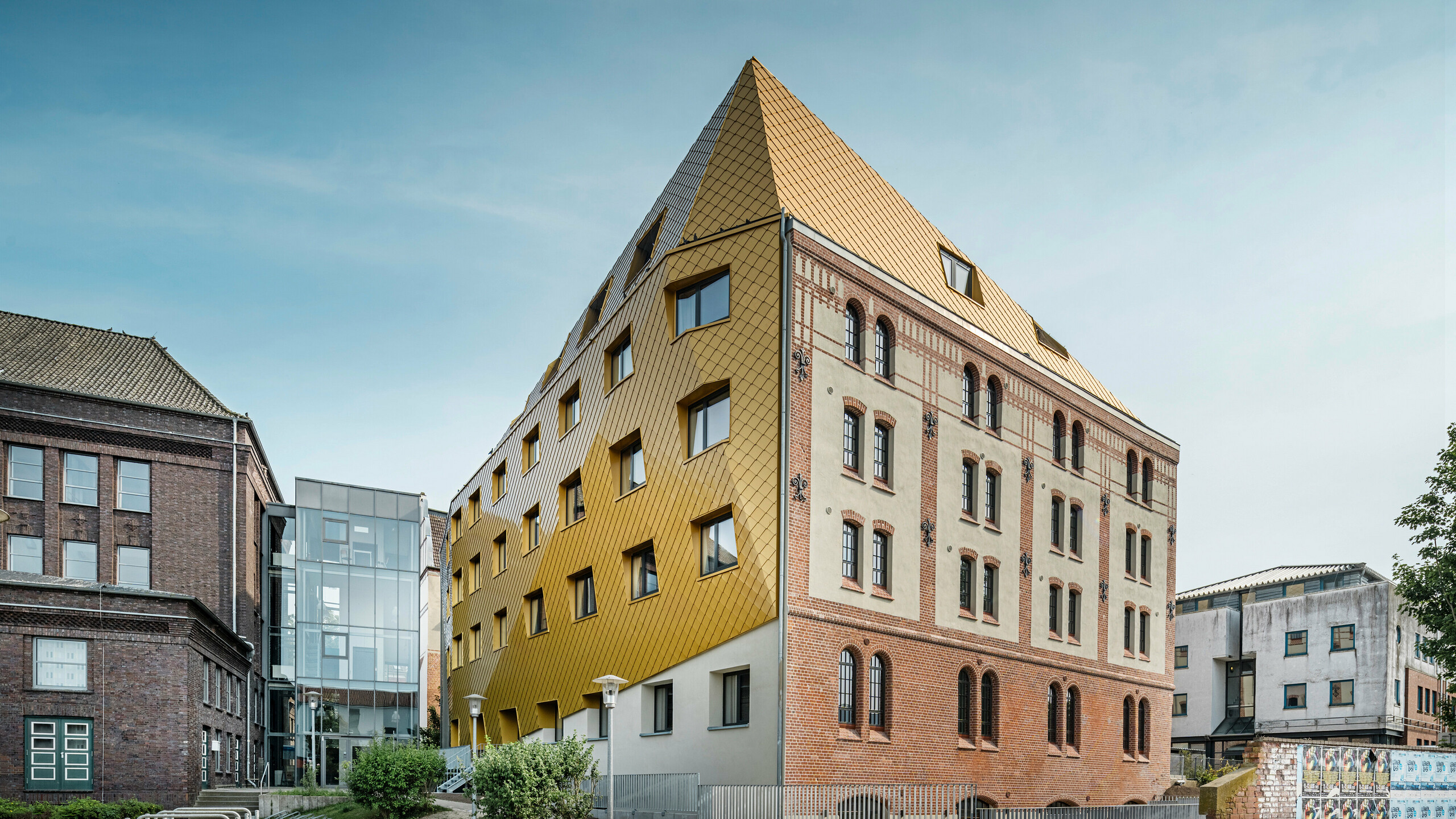 Foto del piano della "Stazione" di Kiel. La residenza per studenti è visibile in diagonale dal lato. L'immagine mostra chiaramente il contrasto tra la facciata in alluminio realizzata con i rombi del tetto e delle pareti PREFA 29 × 29 in Mayagold a sinistra e la facciata in intonaco e mattoni a vista a destra.