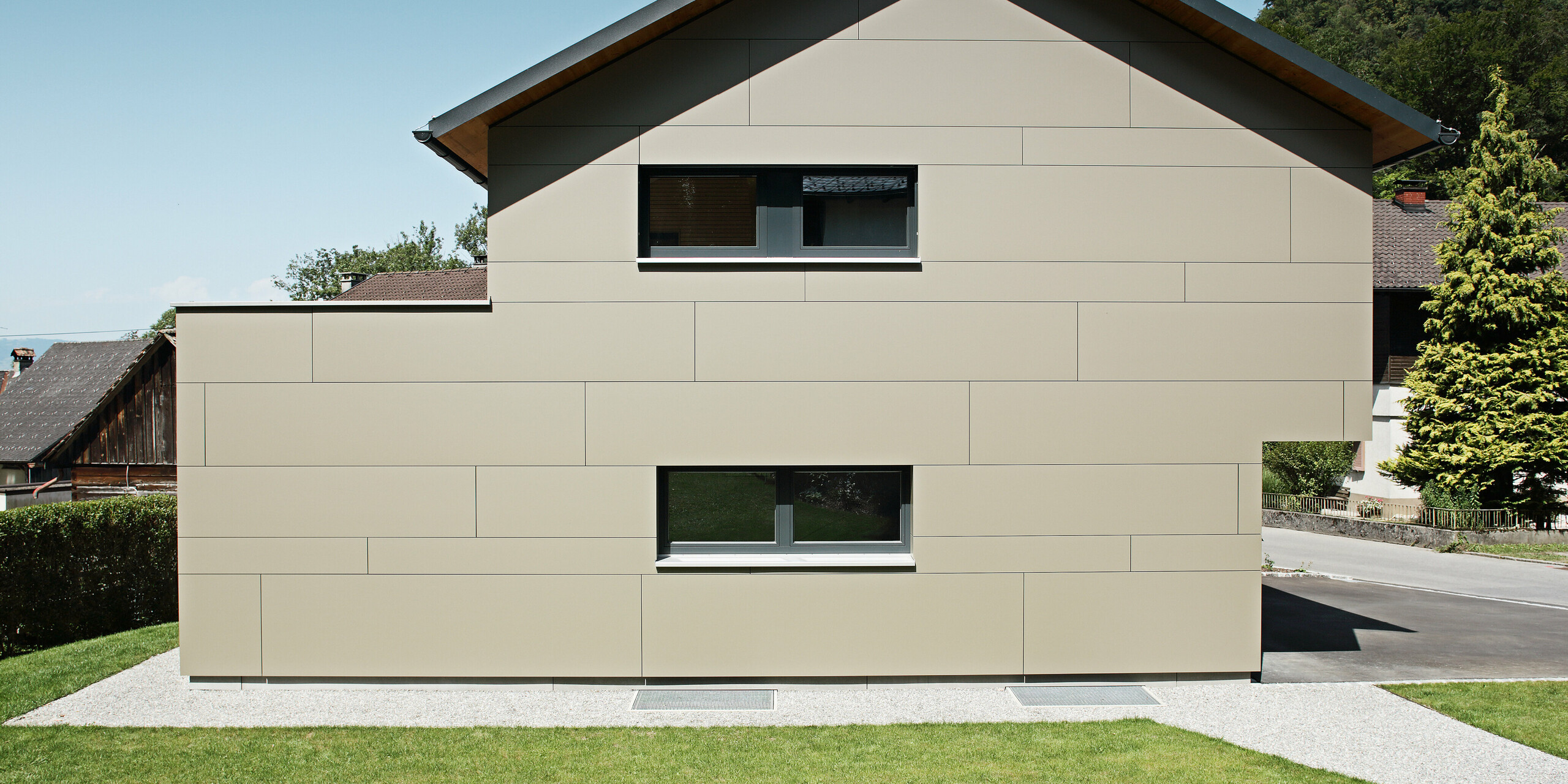 Frontansicht eines modernen Einfamilienhauses in Hohenems, das mit einem PREFA Aluminium Verbundplatten-Fassadensystem in Bronzemetallic verkleidet ist. Die Fassade zeigt eine harmonische Abfolge von horizontalen Linien, die eine ruhige und gleichzeitig zeitgenössische Ästhetik vermitteln. Das Haus fügt sich mit seiner zurückhaltenden Farbgebung und dem klaren Design nahtlos in die umgebende Landschaft ein, während das PREFA Dach – bestehend aus klassischen Dachplatten in P.10 Anthrazit – einen subtilen Kontrast bildet und den langlebigen Charakter des Gebäudes unterstreicht.