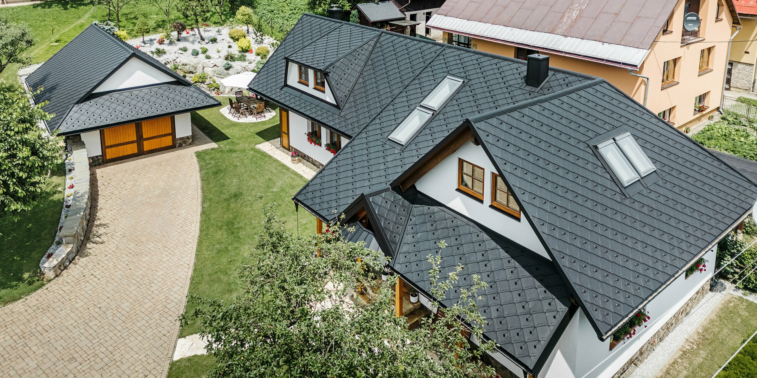 Vogelperspektive auf ein Einfamilienhaus in Stara Bystrica, Slowakei, mit einem Dach aus PREFA Dachrauten 29 × 29 in P.10 Schwarz. Das Dach, ausgeführt in gleichmäßiger Rautenoptik, umfasst Dachschrägenfenster und Trapezgauben auf beiden Seiten. Die Holzverzierungen an den Fenstern sowie die farbenfrohen Blumenarrangements verleihen dem Haus ein einladendes und harmonisches Erscheinungsbild. Der gepflegte Vorgarten und die gepflasterte Einfahrt ergänzen das Gesamtbild perfekt.