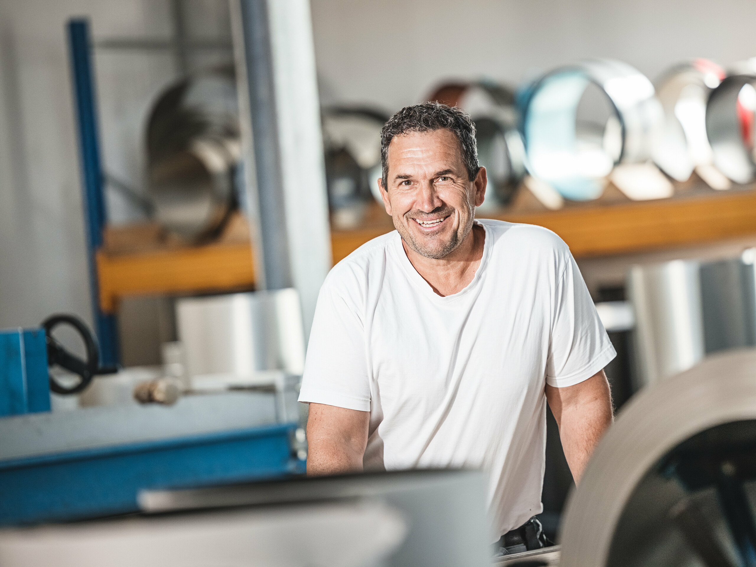 Oskar Urban dell'azienda di coperture Urban nel suo laboratorio. L'esperto installatore PREFA si presenta sorridente davanti a uno sfondo di rotoli di alluminio e macchine che sottolineano l'alta qualità e la precisione del suo lavoro con i prodotti PREFA. La sua esperienza contribuisce in modo significativo alla realizzazione di progetti di successo come il complesso edilizio sul sito di Schmucker a Utting am Ammersee, dove si combinano architettura moderna e materiali sostenibili.