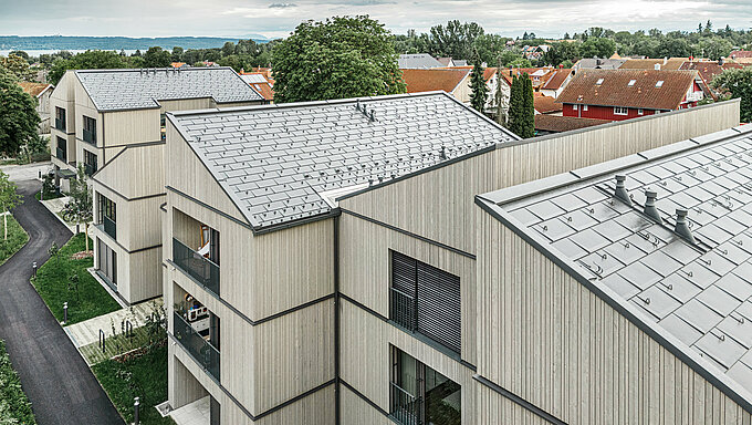 Vista aerea dei moderni condomini di Utting am Ammersee, in Germania. Il progetto residenziale colpisce per l'innovativa costruzione ibrida in legno e per le resistenti tegole R.16 PREFA nel colore P.10 grigio scuro. Gli edifici si inseriscono armoniosamente nel verde circostante e offrono una combinazione estetica di facciate in legno e tetti in alluminio di alta qualità. L'installazione corretta delle tegole garantisce la resistenza agli agenti atmosferici e la durata nel tempo, mentre le linee pulite e l'architettura moderna creano un ambiente di vita invitante e sostenibile. 