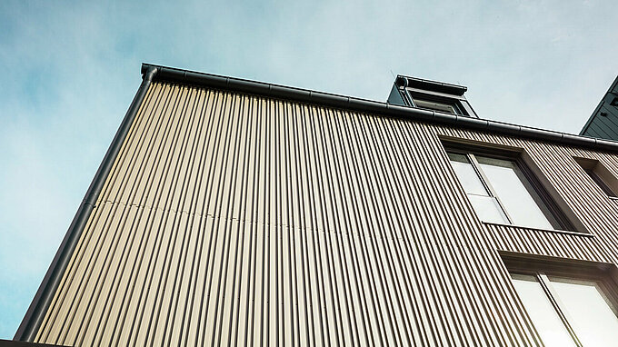 Das Bild zeigt die Fassade eines modernen Gebäudes in Feldkirch, das von Architekt Stefan Heigl entworfen wurde. Die Fassade ist mit Prefalz in der Farbe Bronze verkleidet und verfügt über große Fenster, die viel Tageslicht ins Innere lassen. Die Dachrinne und das Satteldach sind in Schwarzgrau gehalten, ebenfalls mit Prefalz-Materialien. Das elegante und zeitgemäße Design kombiniert hochwertige Materialien und eine funktionale Architektur, die sowohl ästhetisch ansprechend als auch langlebig ist.