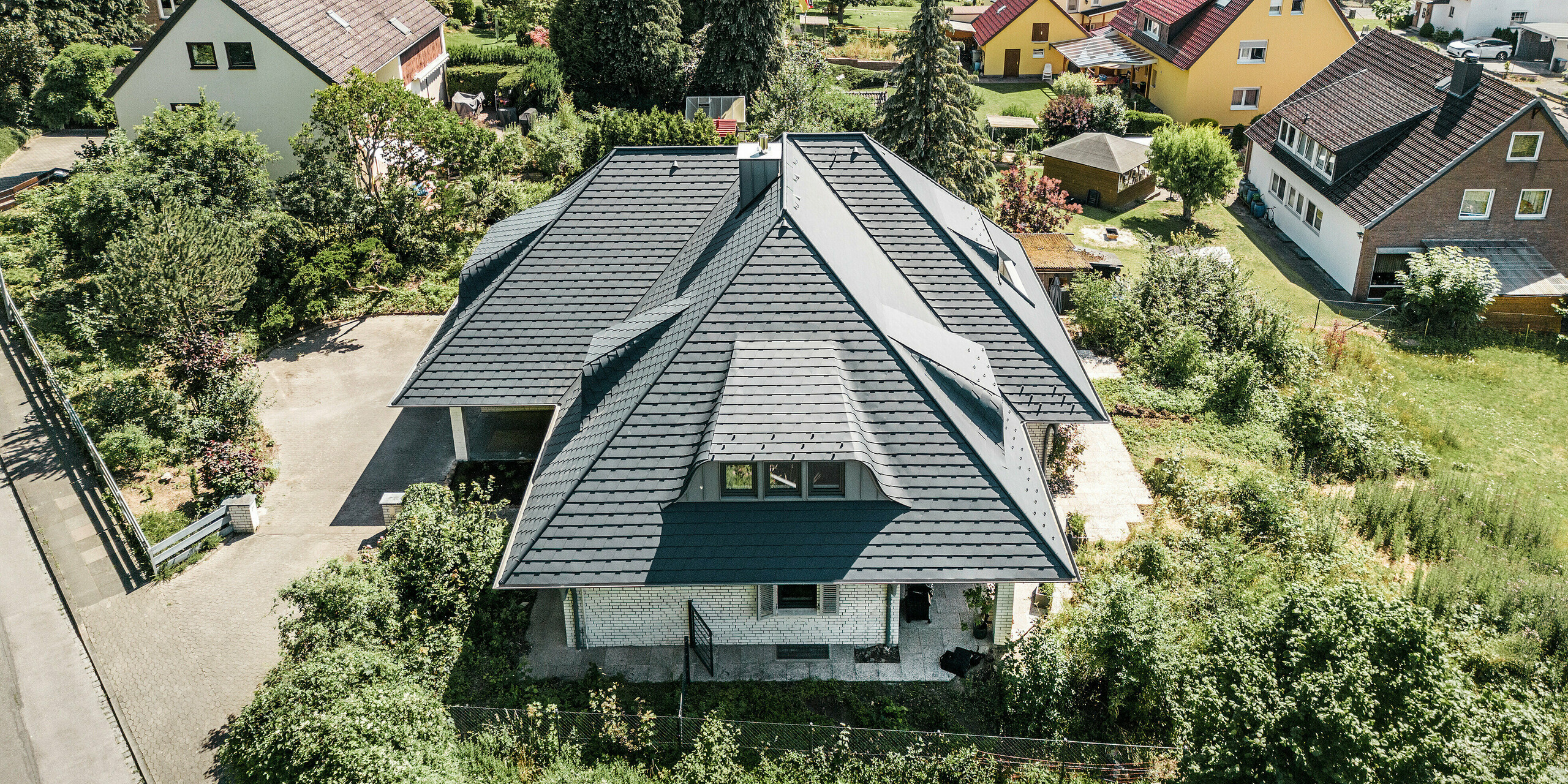 Luftaufnahme eines sanierten Einfamilienhauses in Einbeck mit einem PREFA Dachsystem in P.10 Anthrazit, umgeben von grüner Landschaft. Die Dachschindeln aus Aluminium demonstrieren ihre Vielseitigkeit auf den verschieden geneigten Dachflächen und Kanten, während die präzisen Linien der Dachkanten und Gauben die hochwertige Installation unterstreichen. Die PREFA Dachentwässerung mit PREFA Ablaufrohren und Hängerinnen in Anthrazit fügt sich nahtlos in das Dachdesign ein und bietet eine optimale Funktionalität.