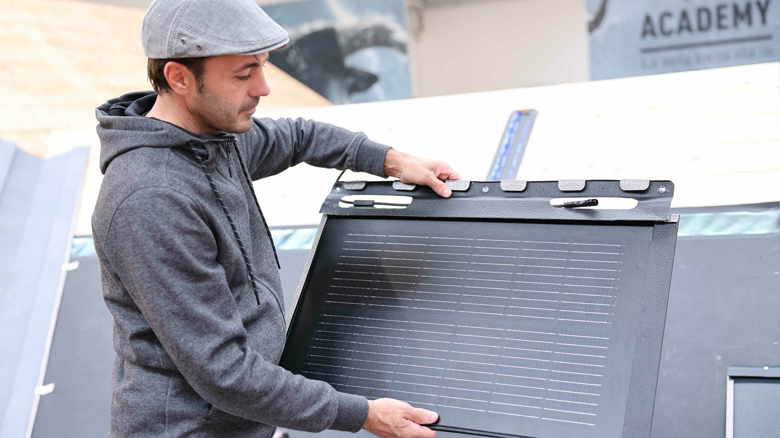 Un dipendente dell'Accademia PREFA in Italia mostra una tegola fotovoltaica da tetto PREFA. Tiene il pannello in mano e ne dimostra il design e le opzioni di montaggio. Sullo sfondo si vedono le aule di formazione dell'Accademia.