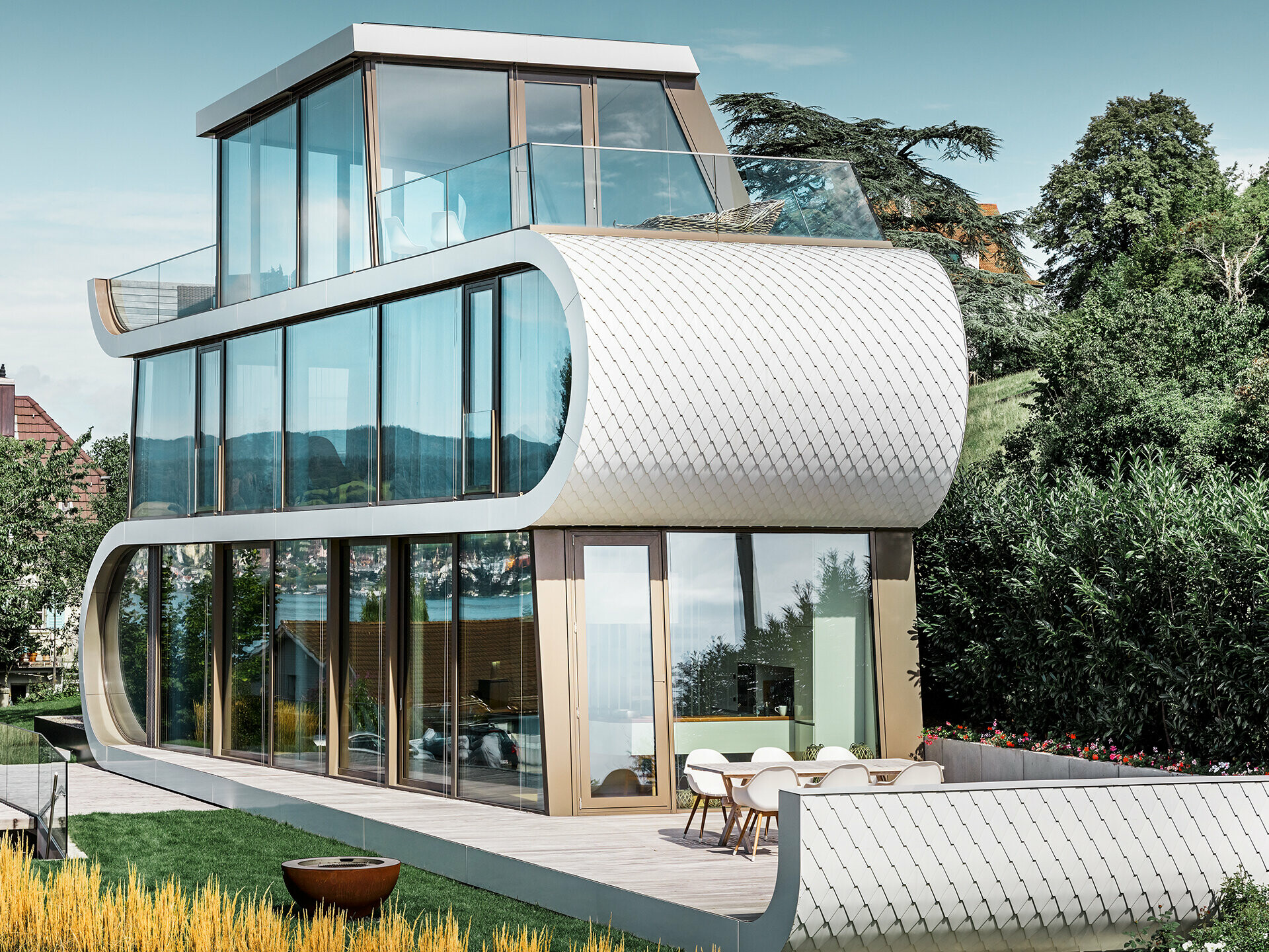 Fotografia del moderno Flexhouse al Zürichsee; con la sua forma insolita il Flexhouse da nell'occhio sempre. La divisione dei piani é realizzate tramite un nastro bianco e curvo che attraversa l'edificio. Le parti curve della facciata sono state rivestite con le scandole in alluminio PREFA nel colore bianco puro