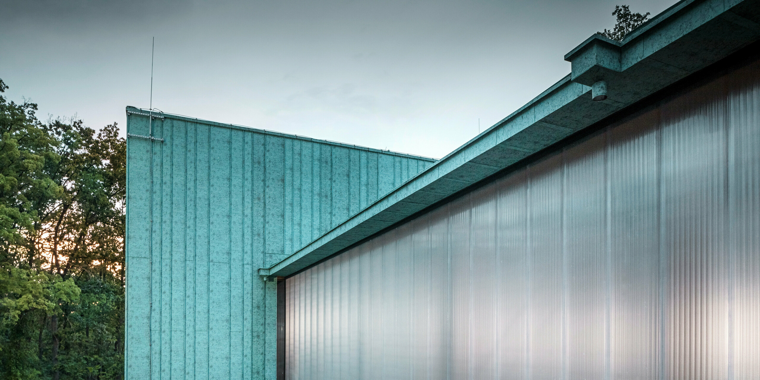 Das Bild zeigt eine seitliche Ansicht des Observatoriums in Fertöboz, Ungarn. Das Gebäude ist mit einer PREFA Aluminiumfassade in Patinagrün verkleidet. Besonders auffällig ist die Kombination aus der lebendigen Fassade und der transluzenten Wand, die im Inneren für eine natürlich Belichtung sorgt. Die Textur und das Farbspiel der Stehfalzverkleidung bieten eine visuelle Tiefe, während die Struktur selbst robust und zeitgemäß erscheint. Im Hintergrund sind Bäume zu sehen, die das natürliche Umfeld betonen, in das das Gebäude eingebettet ist. Der Übergang von der Fassade zum Dach ist nahtlos, was auf eine durchdachte architektonische Planung hinweist. Die Gesamtkomposition ist harmonisch und zeigt, wie moderne Architektur die Balance zwischen Funktionalität, ästhetischer Anziehungskraft und Umweltintegration finden kann.