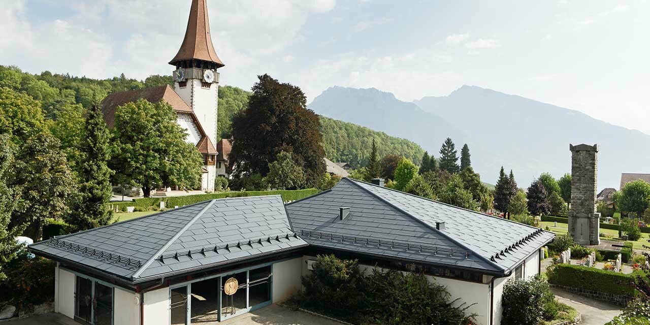 Casa funeraria con pannelli FX.12 PREFA in colore antracite, con una chiesa sullo sfondo e il cimitero accanto.