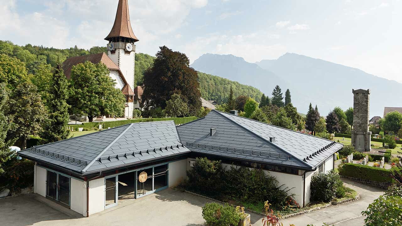 Casa funeraria con pannelli FX.12 PREFA in colore antracite, con una chiesa sullo sfondo e il cimitero accanto.