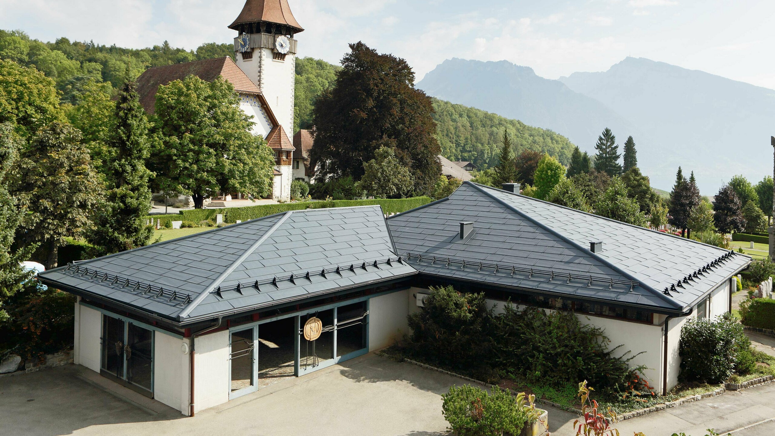 Aufbahrungshalle mit FX.12 Dachpaneelen von PREFA in Anthrazit, im Hintergrund steht eine Kirche und daneben befindet sich der Friedhof.